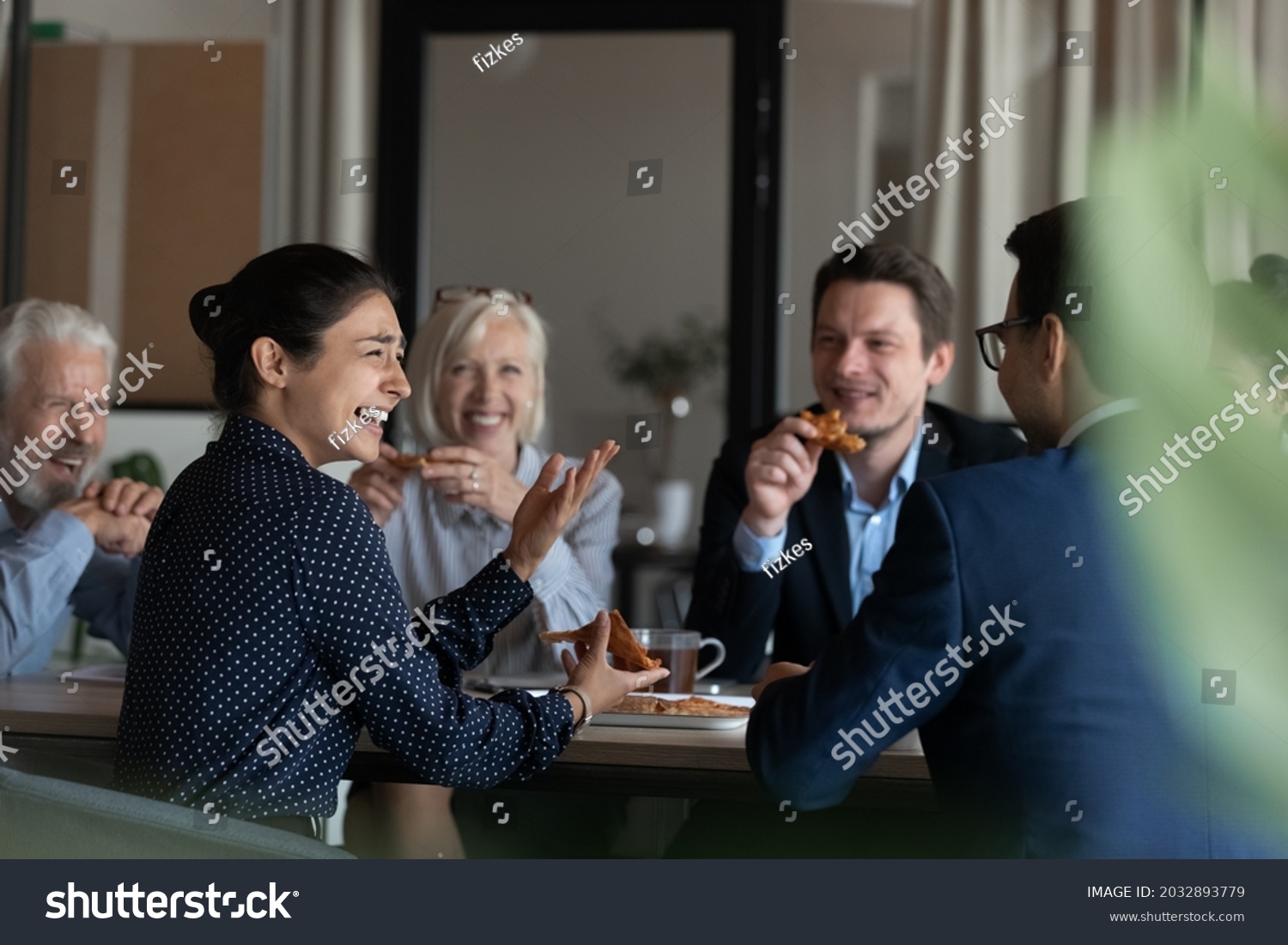 Excited diverse employees eating pizza during break in office together, happy Indian businesswoman laughing at funny joke, talking chatting with colleagues, having fun, sharing corporate lunch #2032893779