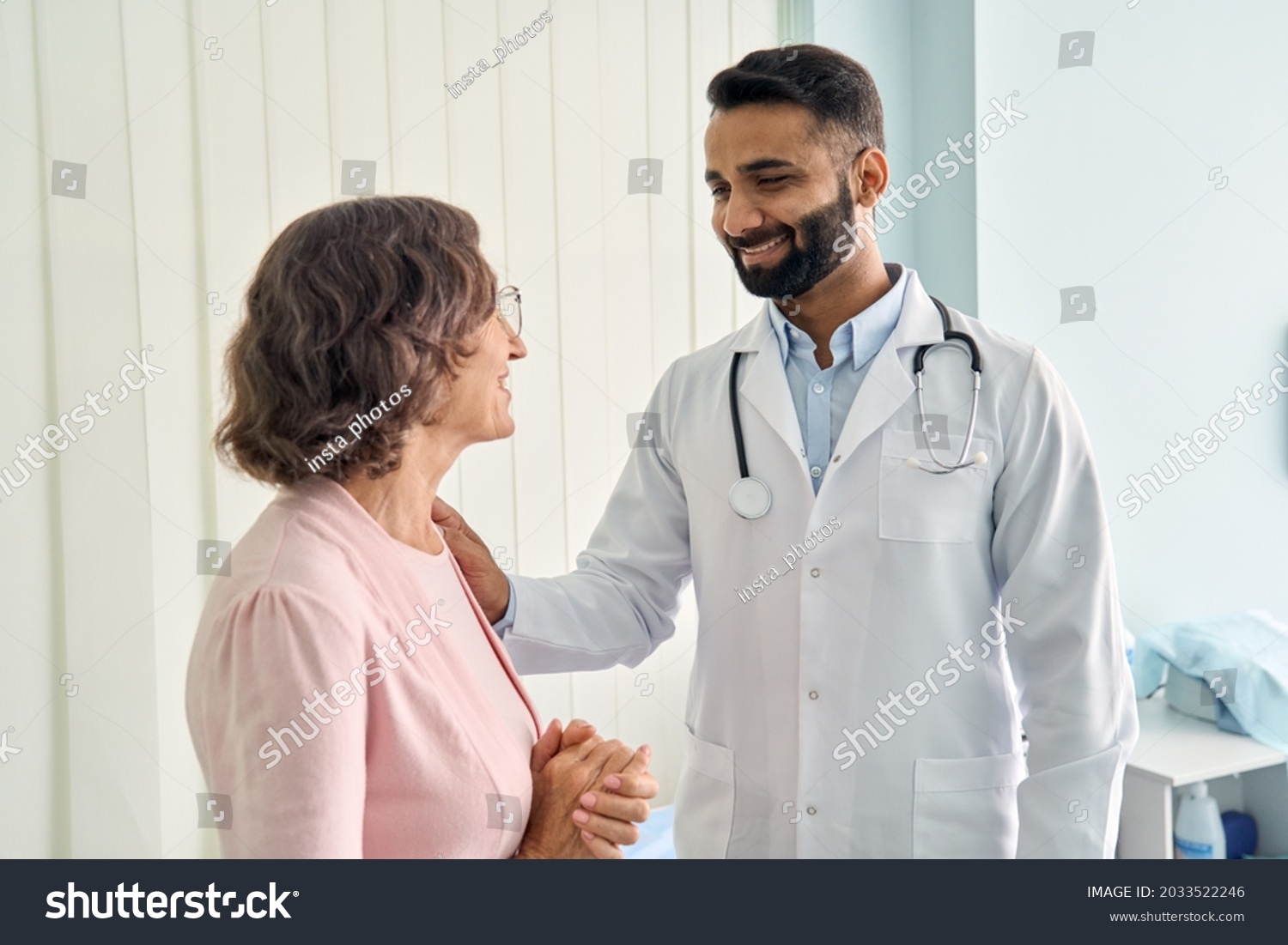 Happy young Indian doctor therapist in white coat has appointment consulting supporting putting hand on shoulder of older senior female patient in modern clinic hospital. Medical healthcare concept. #2033522246