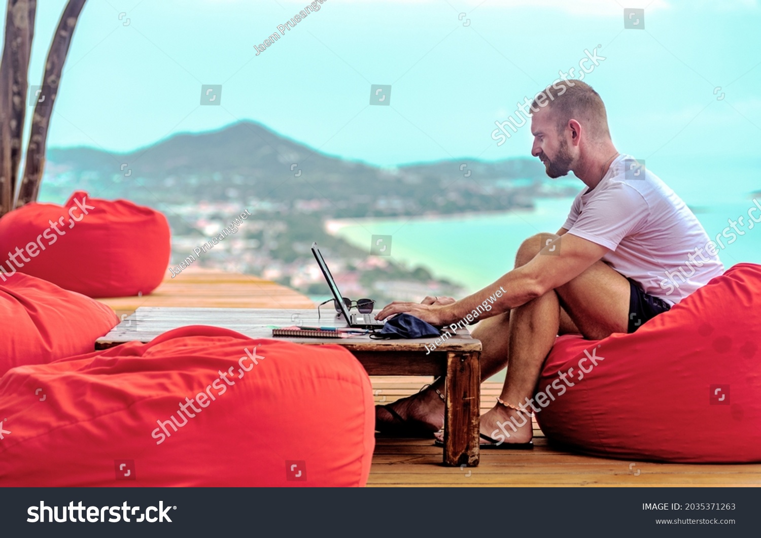 A Caucasian man sitting on a red cushion working remotely with his laptop. There are mountains and the sea in the background #2035371263