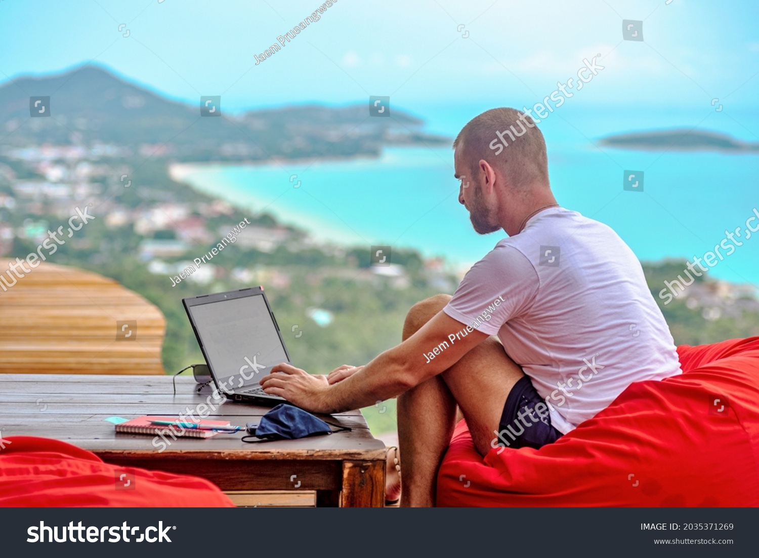 A Caucasian man sitting on a red cushion working remotely with his laptop. There are mountains and the sea in the background #2035371269
