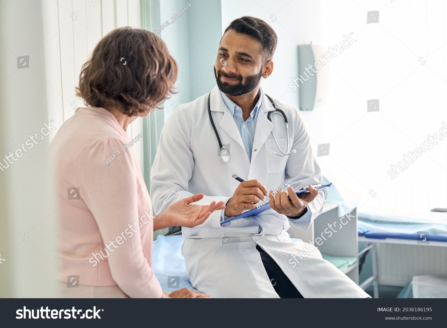Indian male doctor consulting senior old patient filling form at consultation. Professional physician wearing white coat talking to mature woman signing medical paper at appointment visit in clinic. #2036186195