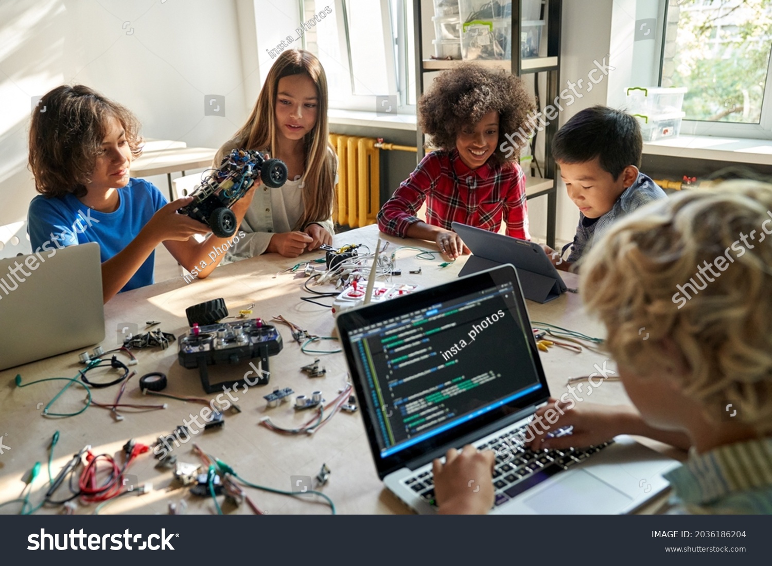 Diverse school children students build robotic cars using computers and coding. Happy multiethnic kids learning programming robot vehicles sitting at table at STEM education science engineering class. #2036186204