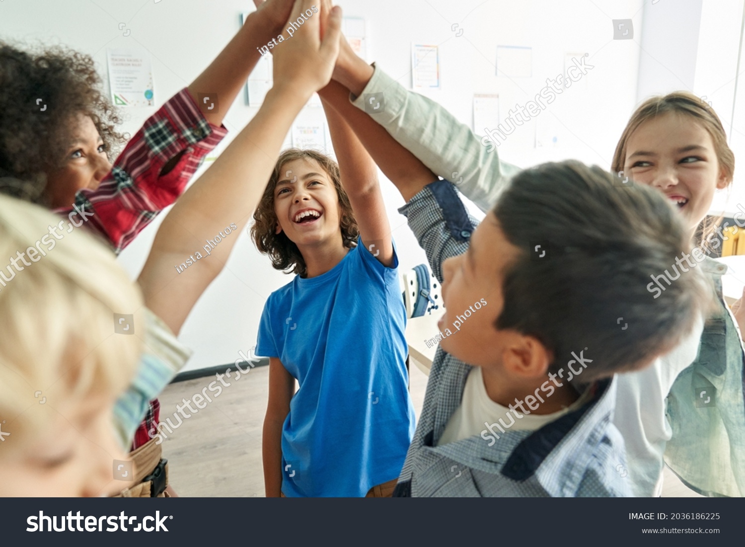 Happy diverse multiethnic kids junior school students group giving high five together in classroom. Excited children celebrating achievements, teamwork, diversity and friendship with highfive concept. #2036186225