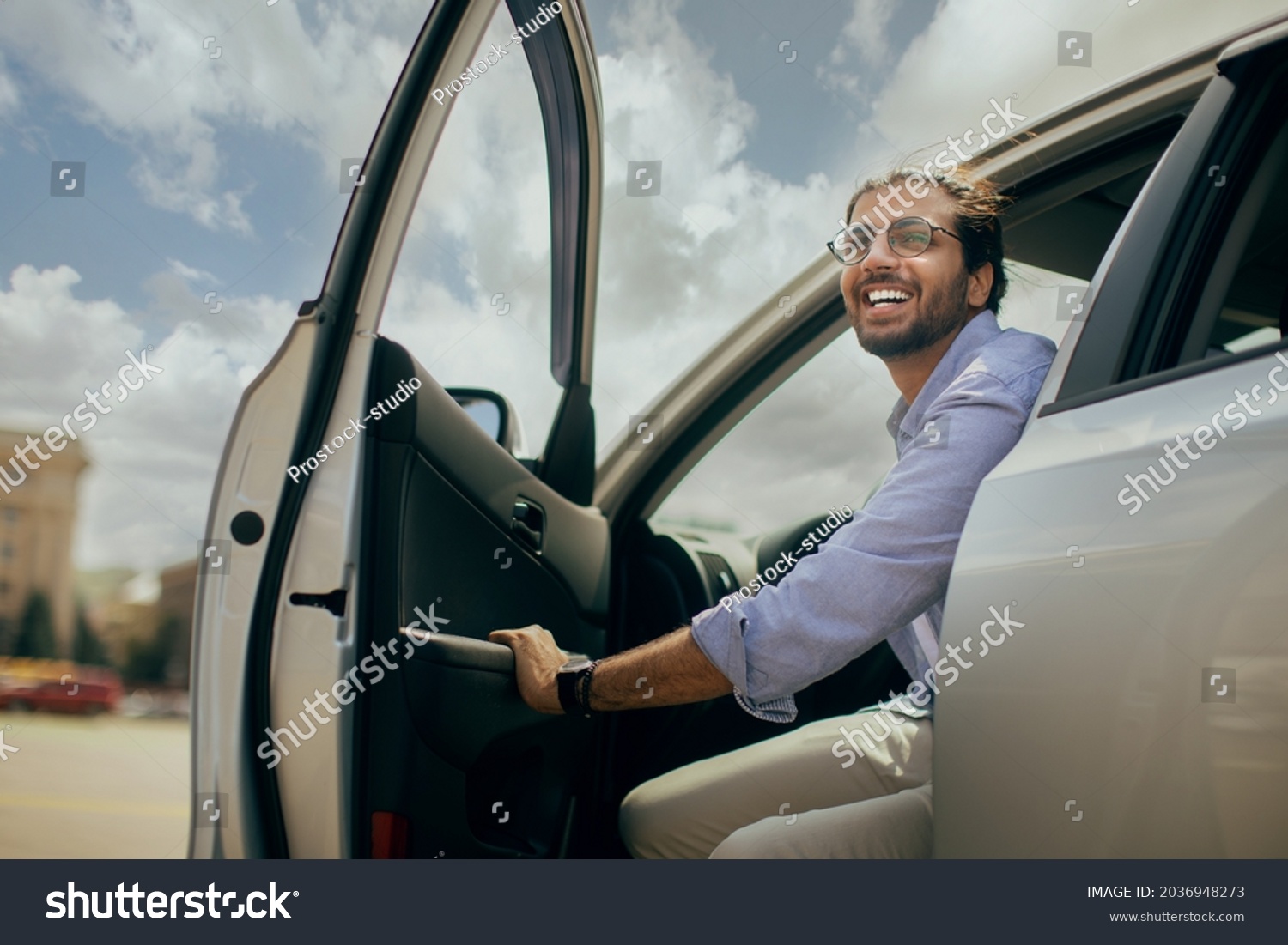 Positive young middle-eastern man going out the car, low angle photo, copy space. Happy arab guy open automobile door and smiling, getting in. Auto test-drive, leasing, buying, renting concept #2036948273