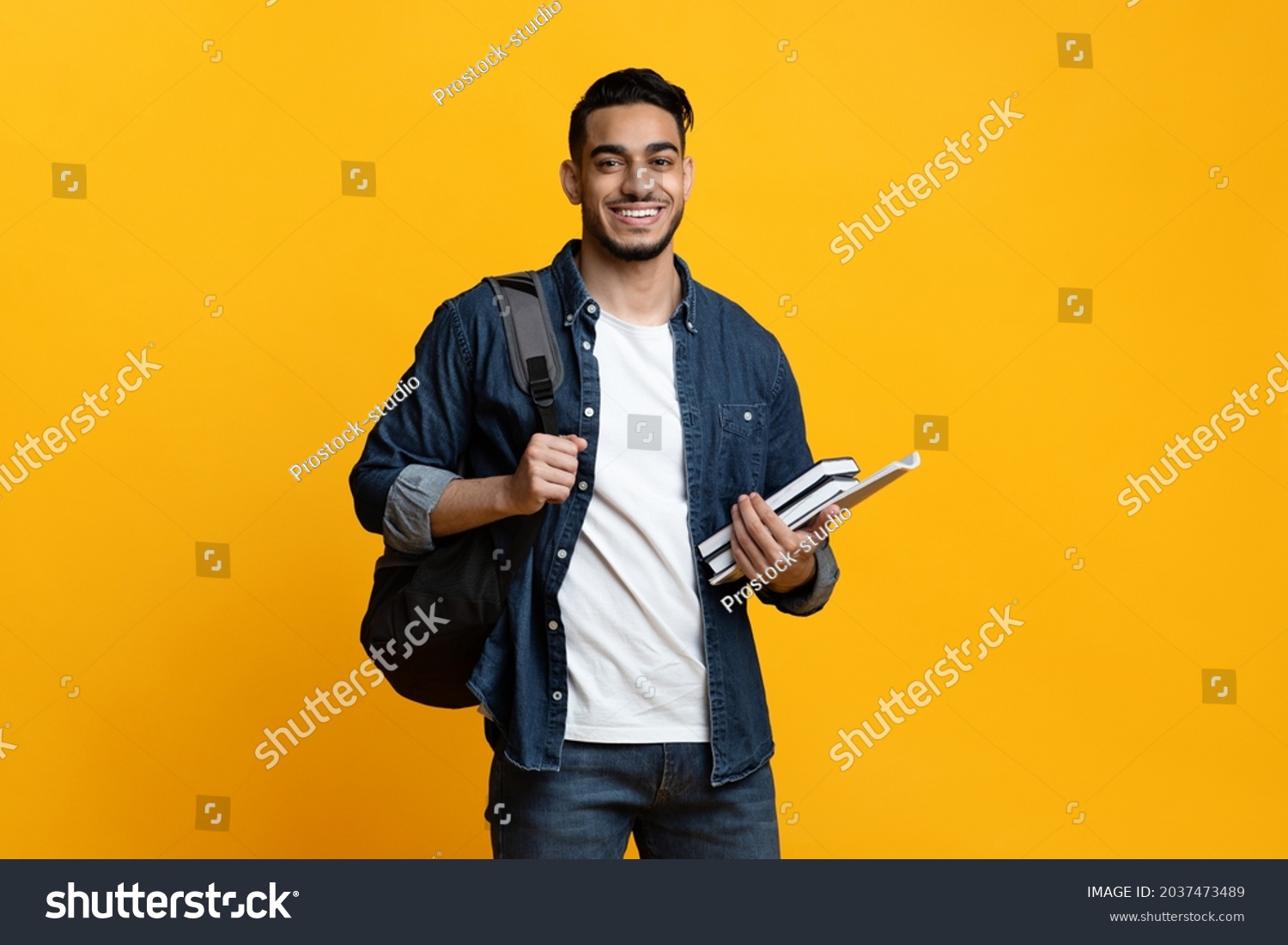 Smart arab guy student with backpack and bunch of books smiling at camera, copy space for advertisement over yellow studio background. Education, university, college, studying, course concept #2037473489
