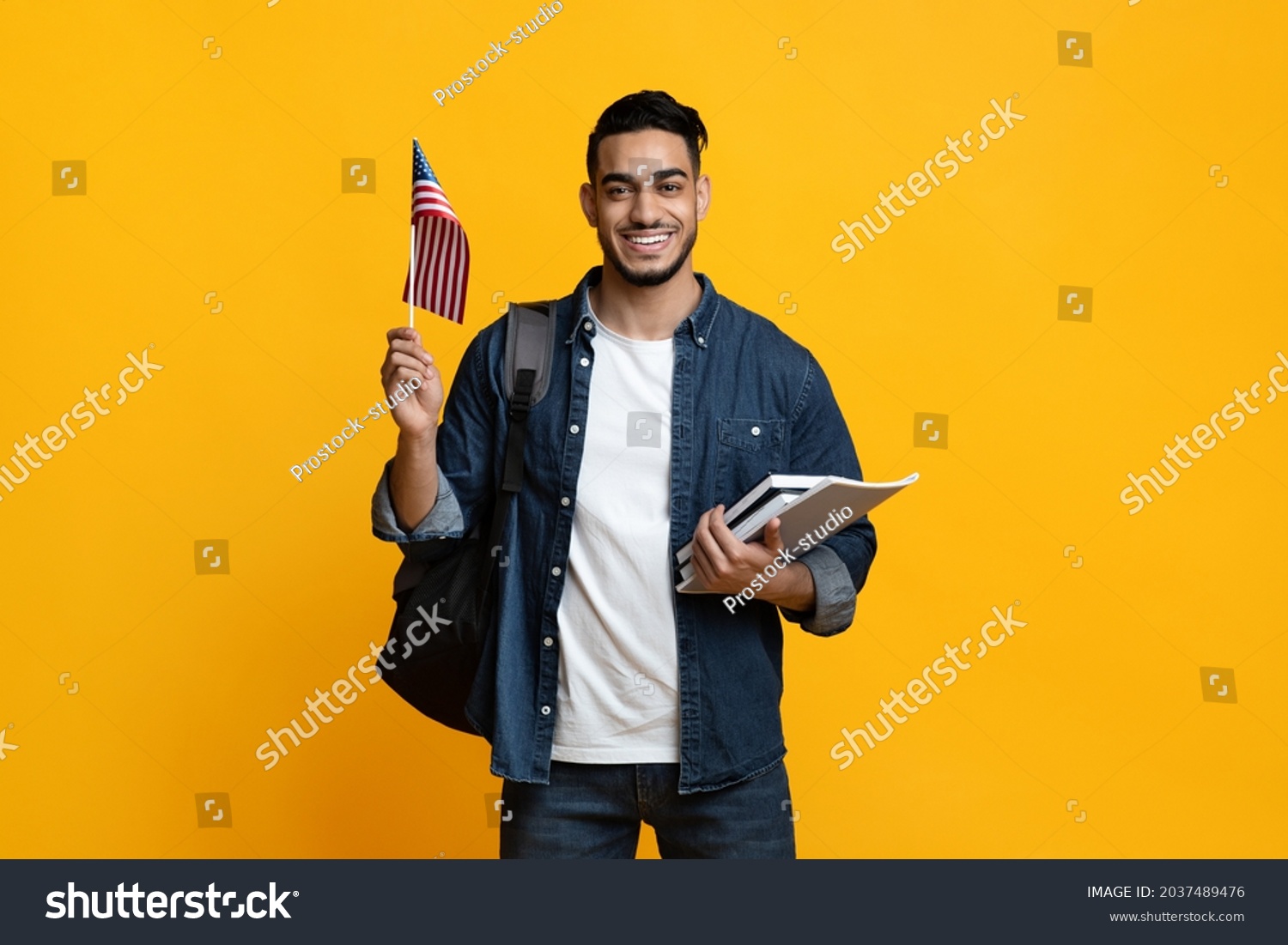 Smiling arab guy student with backpack and bunch of books showing flag of the US over yellow studio background, copy space. Positive middle-eastern young man studying English at school #2037489476