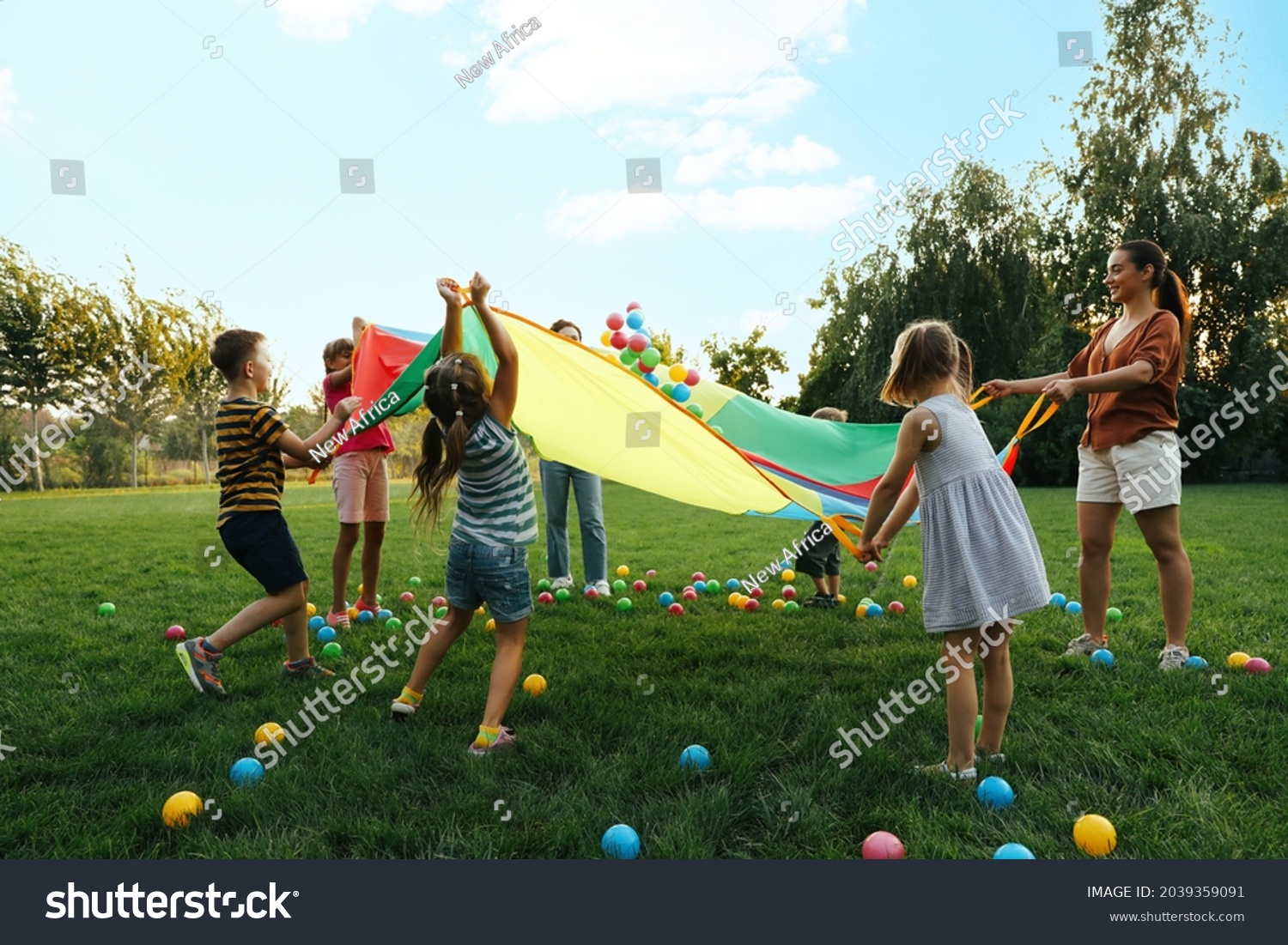 Group of children and teacher playing with rainbow playground parachute on green grass. Summer camp activity #2039359091