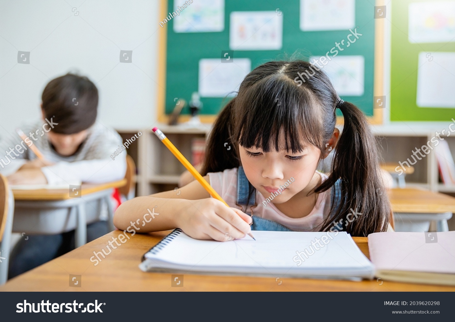 Portrait of little pupil writing at desk in classroom at the elementary school. Student girl study doing test in primary school. Children writing notes in classroom. Education knowledge concept #2039620298