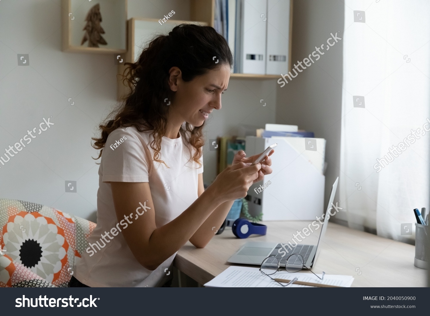 Irritated stressed woman sit at desk hold cellphone look at screen feels annoyed due spam, missed call, crashed app, bad business news, problems with not working, discharged or broken device concept #2040050900