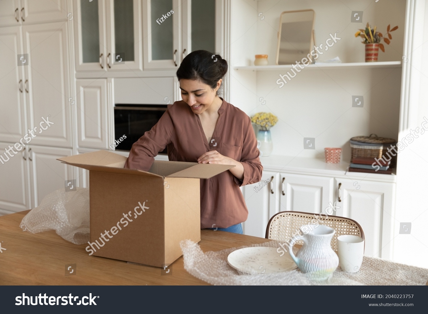 Just after relocation. Happy biracial woman unpack stuff take ceramic tableware out of big carton container at modern kitchen. Young indian lady get internet purchase open box with newly bought dishes #2040223757