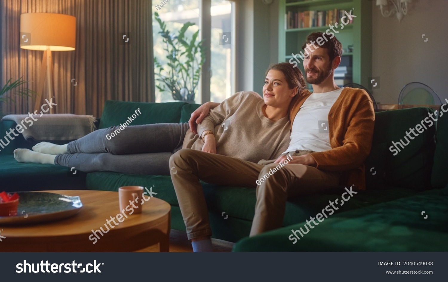 Couple Watches TV together while Sitting on a Couch in the Living Room. Girlfriend and Boyfriend embrace, cuddle, talk, smile and watch Television Streaming Services. Home with Cozy Stylish Interior. #2040549038