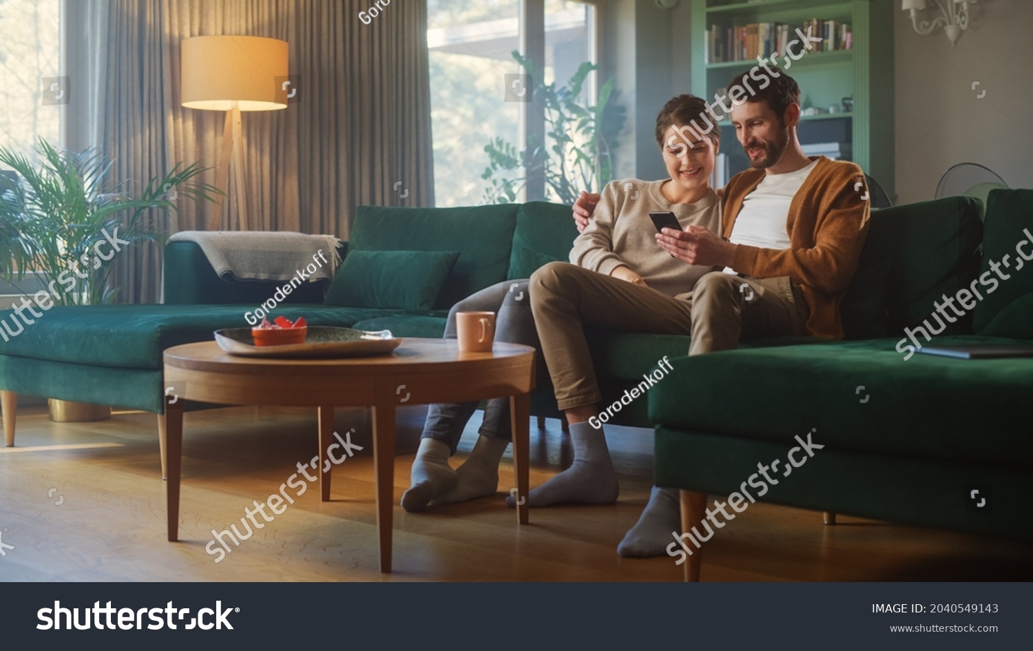 Couple Use Smartphone Device, while Sitting on a Couch in the Cozy Apartment. Boyfriend and Girlfriend Talk, do e-Shopping on Internet, Watching Funny Videos, Use Social Media, Streaming Service. #2040549143