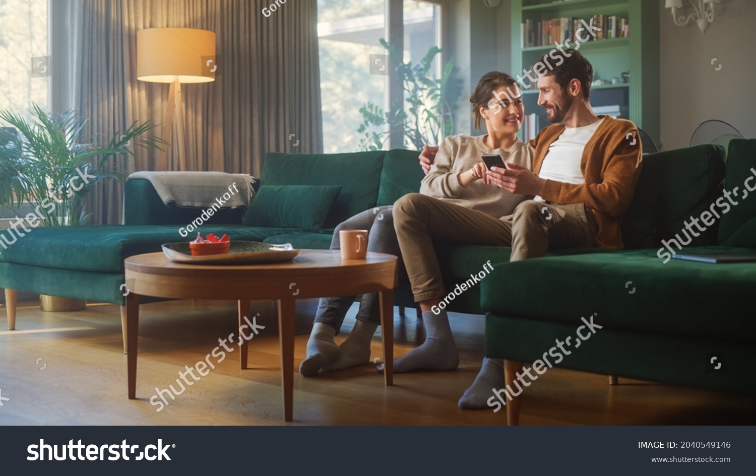 Couple Use Smartphone Device, while Sitting on a Couch in the Cozy Apartment. Boyfriend and Girlfriend Talk, do e-Shopping on Internet, Watching Funny Videos, Use Social Media, Streaming Service. #2040549146