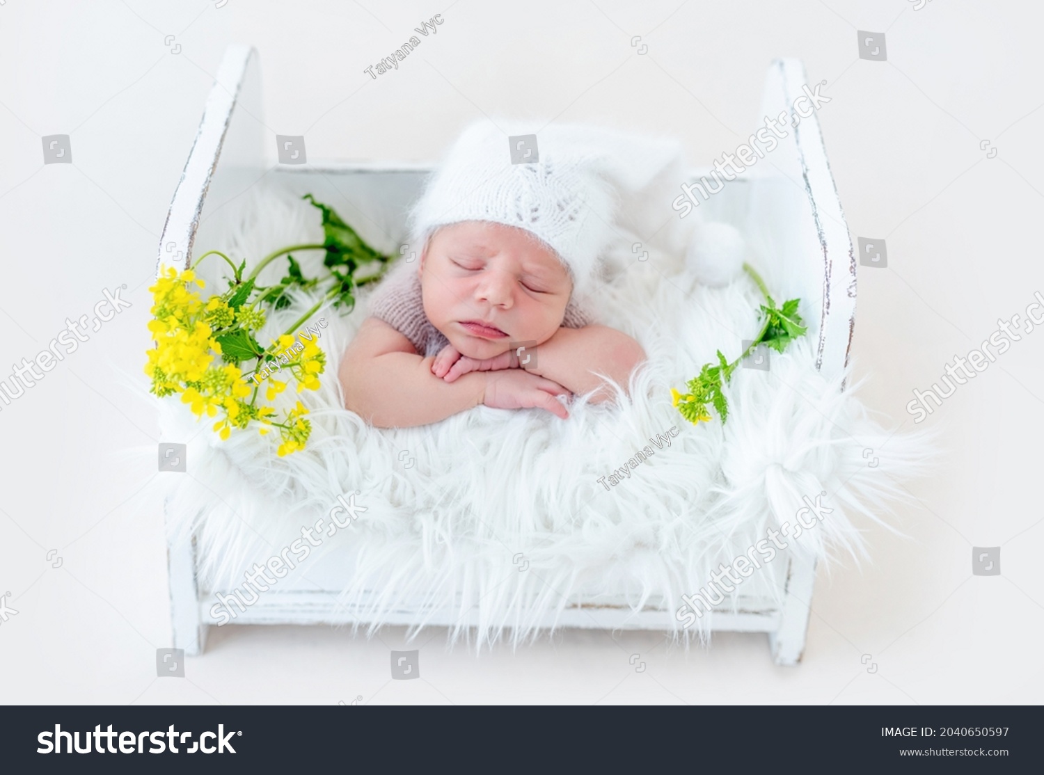 Adorable newborn baby boy sleeping on his tummy in white wooden bed with yellow flowers. Cute infant kid wearing knitted hat napping during studio photoshoot #2040650597
