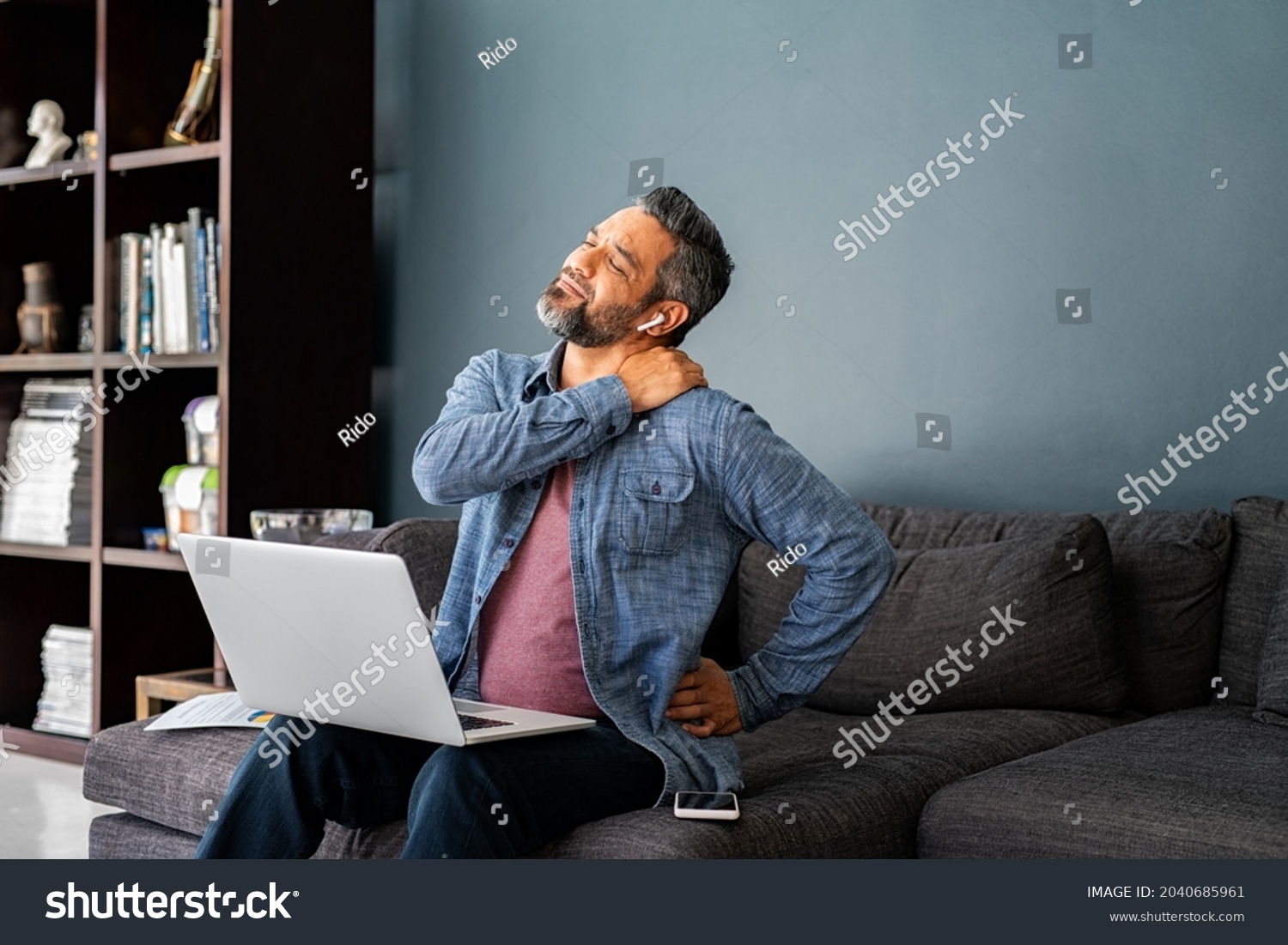 Mature indian man suffering from shoulder and back pain while sitting on couch and working from home on laptop. Stressed middle eastern businessman suffering from neck pain and stretching. #2040685961
