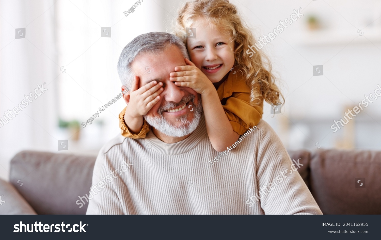 Cute little girl covering eyes with hands of her smiling senior grandfather while playing and having fun together at home, small happy child spending time with positive active grandpa in living room #2041162955