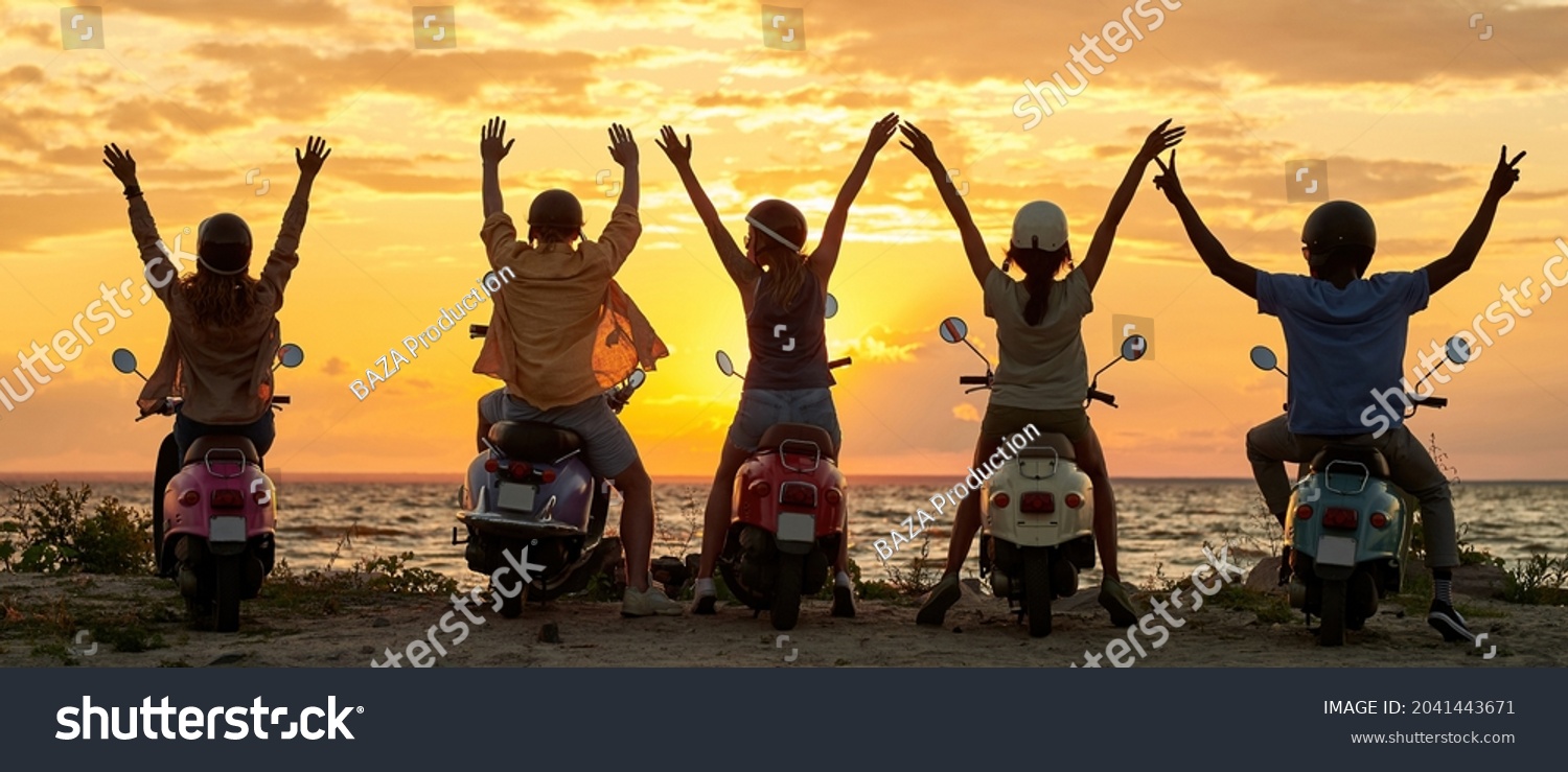 Back view of cheerful young friends, motorcycle riders raised their hands while standing with their scooters on the beach and watching sunset together. Friendship, nature concept. Rear view #2041443671