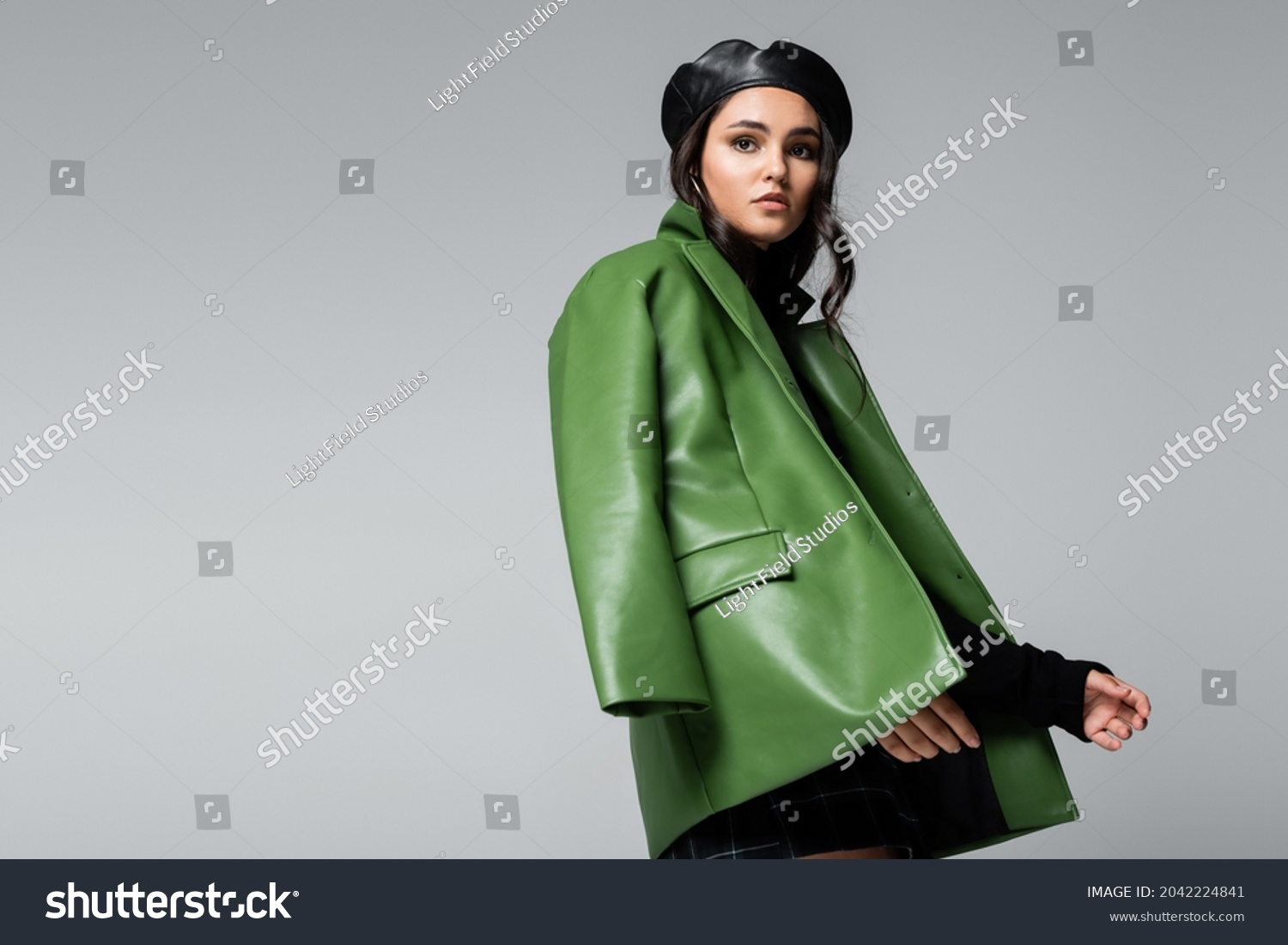 young trendy woman in black beret and green leather jacket posing isolated on grey #2042224841