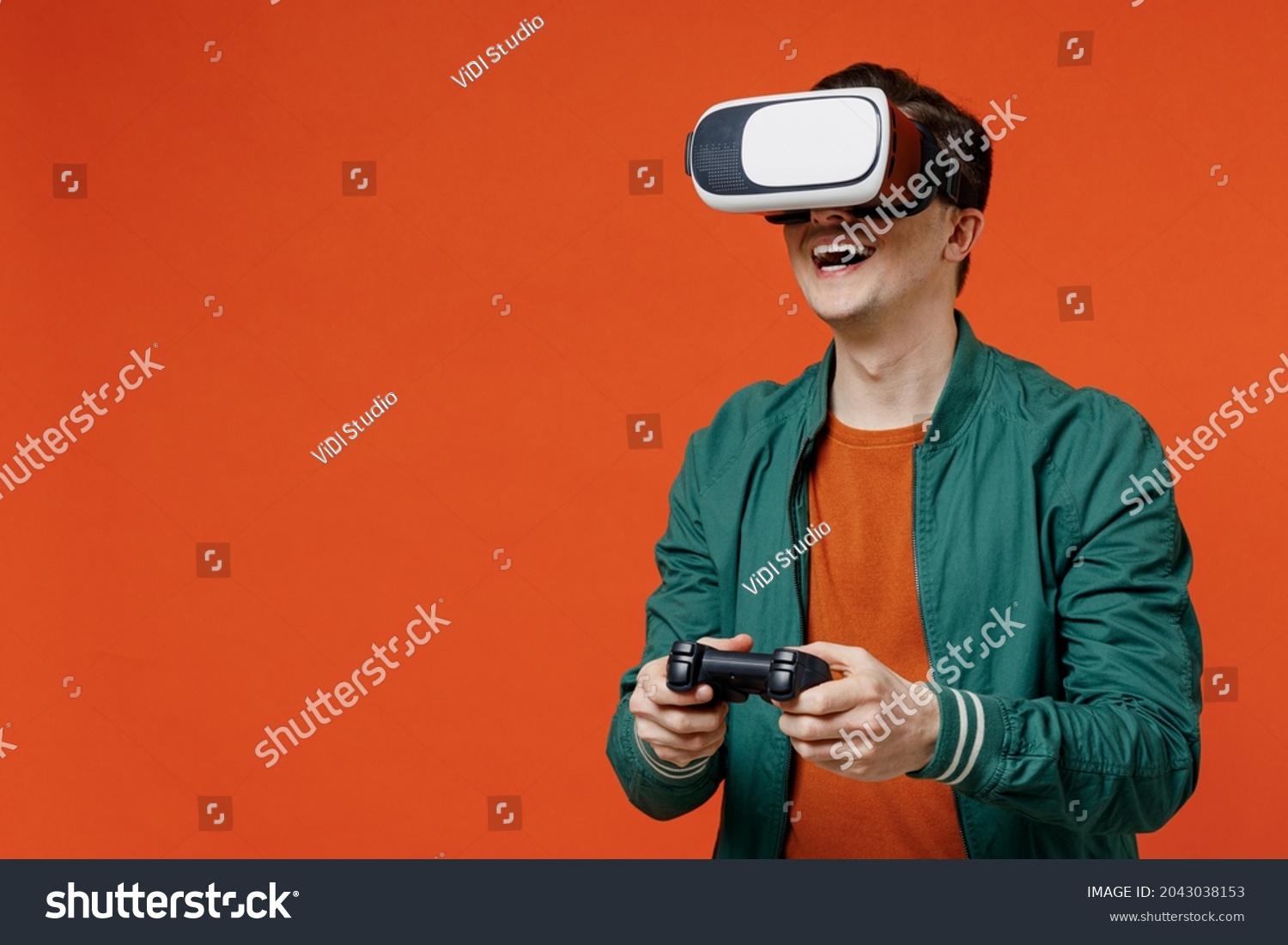 Smiling vivid excited happy young brunet man 20s wear red t-shirt green jacket watching in vr headset pc gadget play pc game with joystick console isolated on plain orange background studio portrait. #2043038153