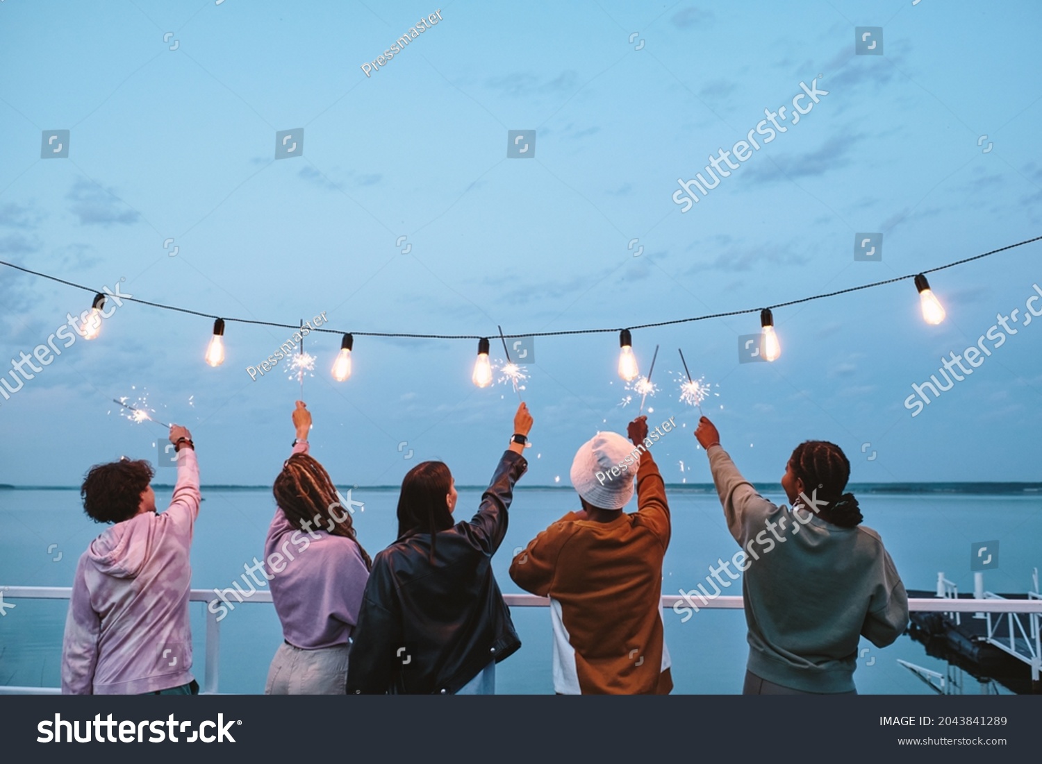 Back view of young ecstatic friends with bengal lights enjoying summer evening by lake #2043841289
