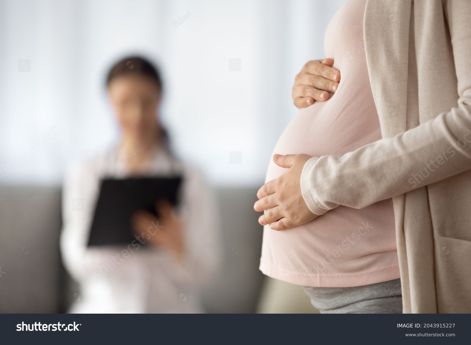 Preparing for baby birth. Close up of woman expecting baby having appointment with doctor at antenatal clinic prenatal healthcare center. Focus on young female holding hands on baby bump in doc office #2043915227