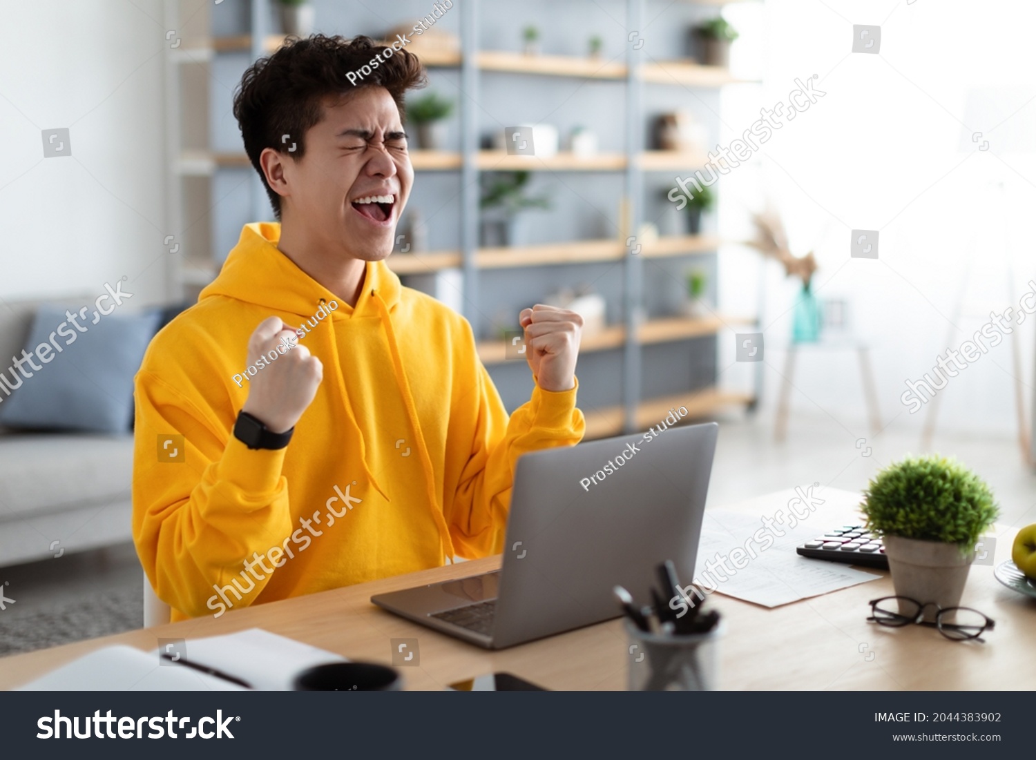Excited asian man emotionally celebrating success using laptop in home office, sitting at desk raising hands with joy, shaking fists, received good news, got promotion, finished project, free space #2044383902