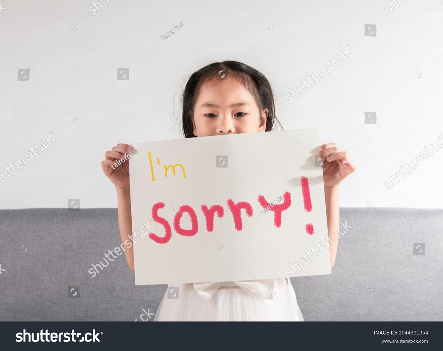 Cute Asian kid little toddler girl feels guilty is holding a sign of "I'm sorry" written on a white paper to apologize to her mother, father, or teacher for her mistake at home or school. #2044391954