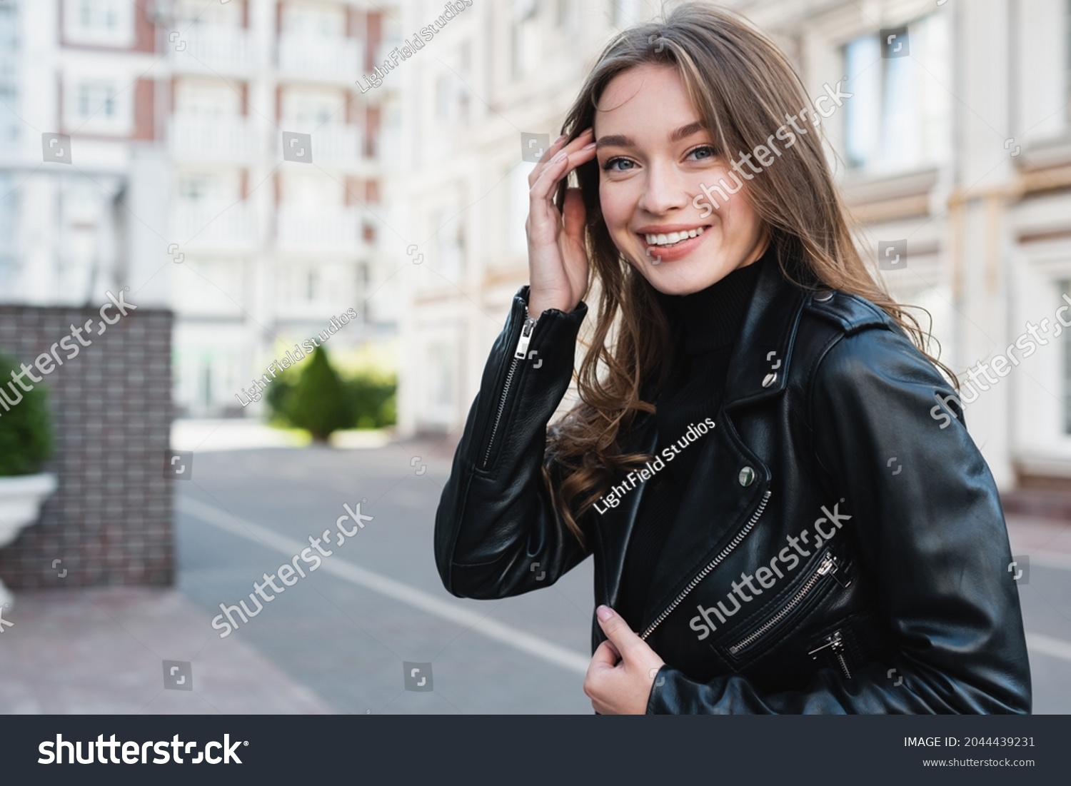 happy young woman in black turtleneck and leather jacket adjusting hair on urban street of europe #2044439231