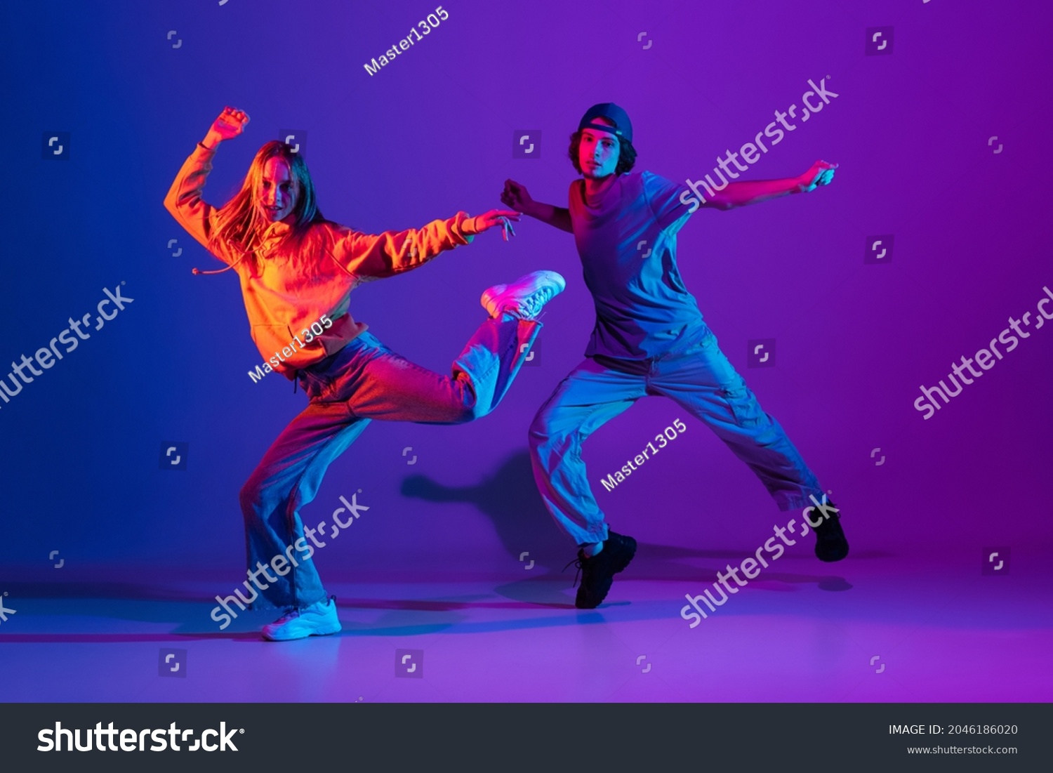 Stylish man and woman dancing hip-hop in casual sports youth clothes on gradient purple pink background at dance hall in neon light. Youth culture, hip-hop, movement, style and fashion, action. #2046186020