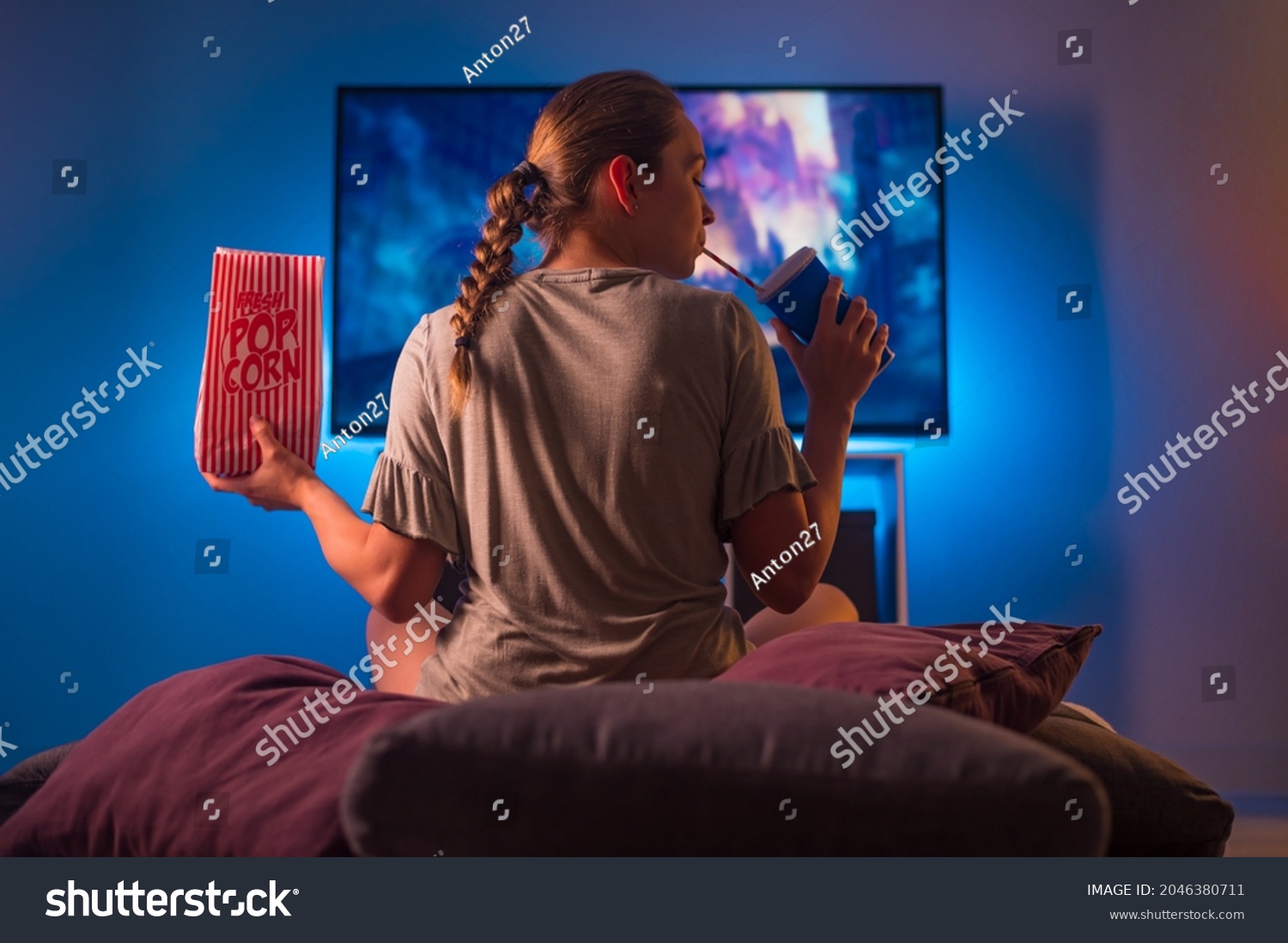 A blonde girl in pajamas watches TV at night. Insomnia. She is holding a pack of popcorn and a carbonated drink with a straw. Neon light. Watching TV shows and series, quarantine. #2046380711