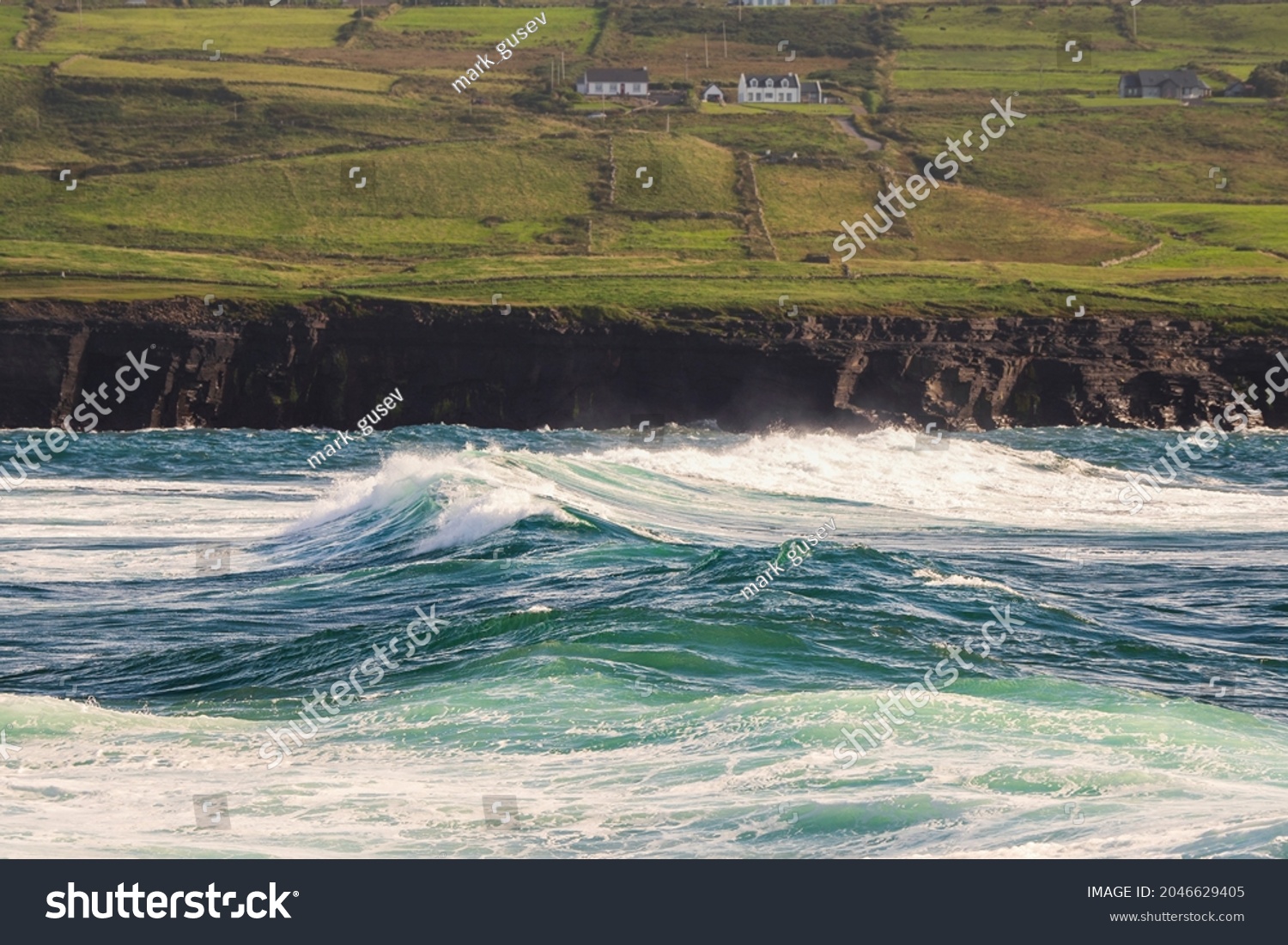 Powerful waves crushing against Cliffs and rough stone coastline of West coast of Ireland. Doolin area. County Clare. Ocean power and rugged Irish coastline. Green fields in the background #2046629405