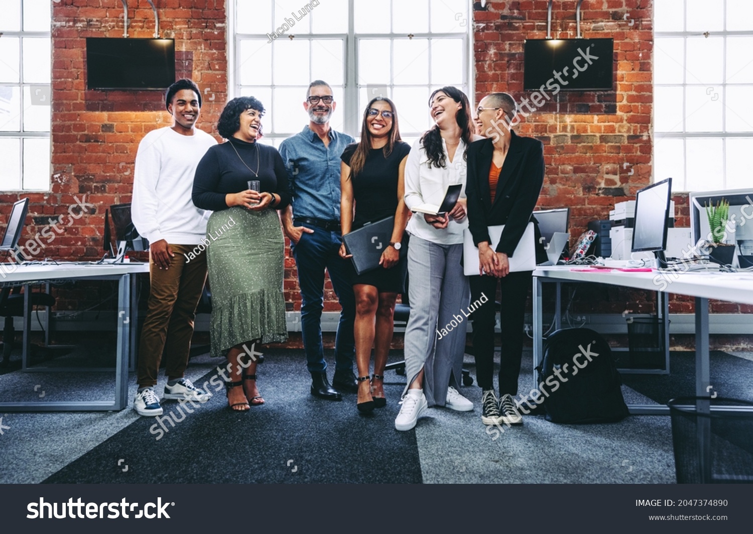Happy group of businesspeople laughing cheerfully in a modern workplace. Diverse group of colleagues enjoying working together in an office. Successful businesspeople standing together. #2047374890