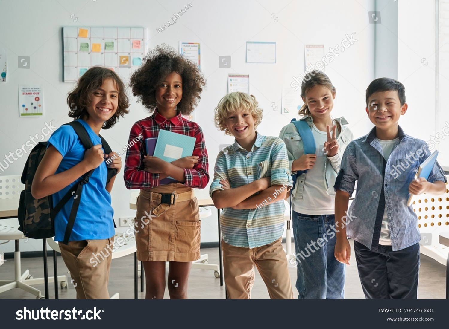 Portrait of cheerful smiling diverse schoolchildren standing posing in classroom holding notebooks and backpacks looking at camera happy after school reopen. Diversity. Back to school concept. #2047463681