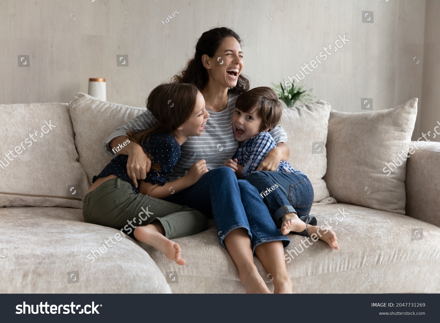 Overjoyed young hispanic mother cuddling small laughing kids siblings, having fun entertaining resting together on comfortable sofa. Joyful multigenerational family playing on weekend at home. #2047731269