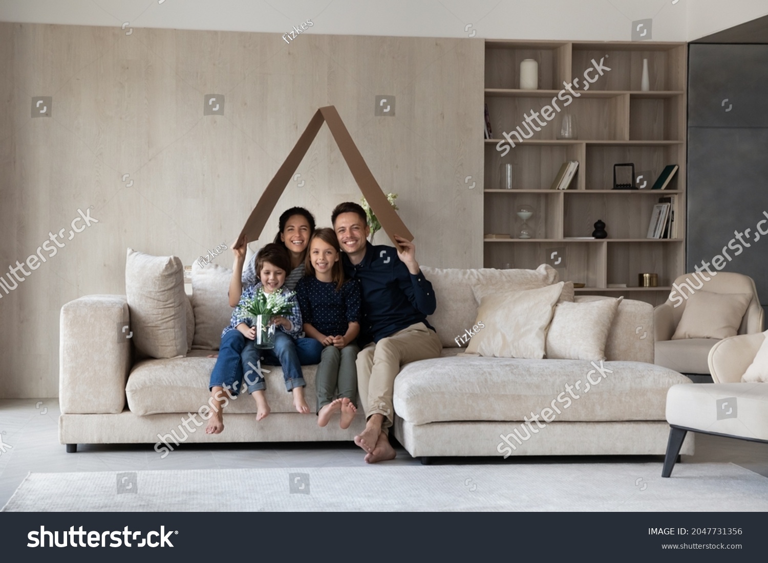 Full length joyful young married hispanic couple parents sitting with little children son daughter on comfortable couch under carton roof, celebrating moving into own dwelling, real estate concept. #2047731356