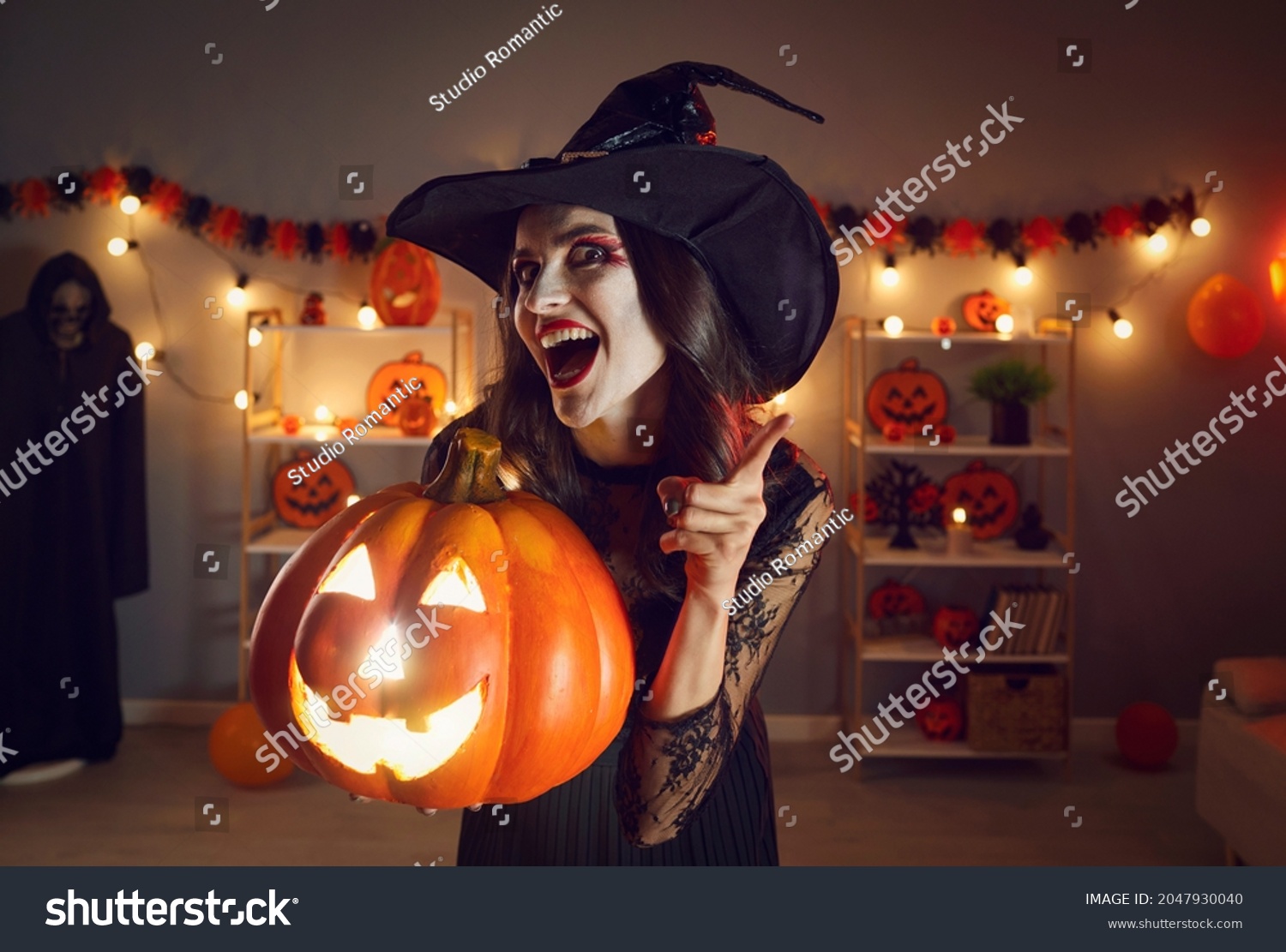 Portrait of woman in wicked witch costume. Beautiful lady in black pointed hat with happy excited face expression standing in dark room, holding Halloween jack-o-lantern pumpkin, shaking finger at you #2047930040