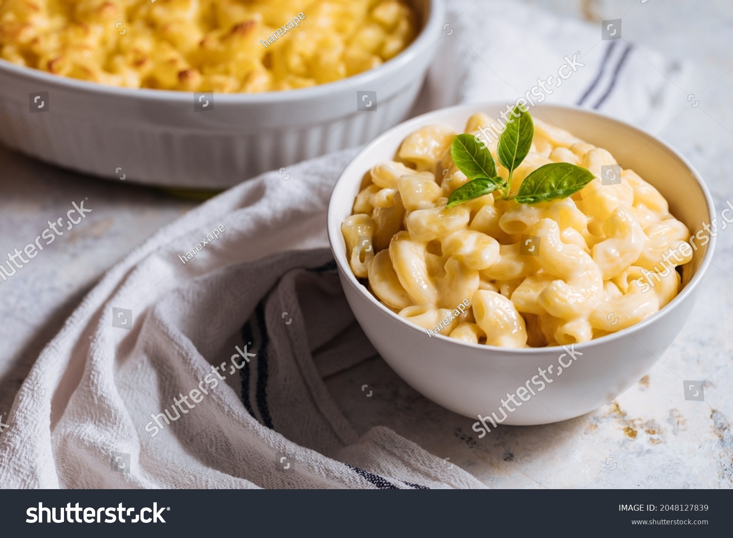 Mac and cheese in white bowl with basil on top and another mac and cheese baked in oven in background placed on a white rustic board #2048127839