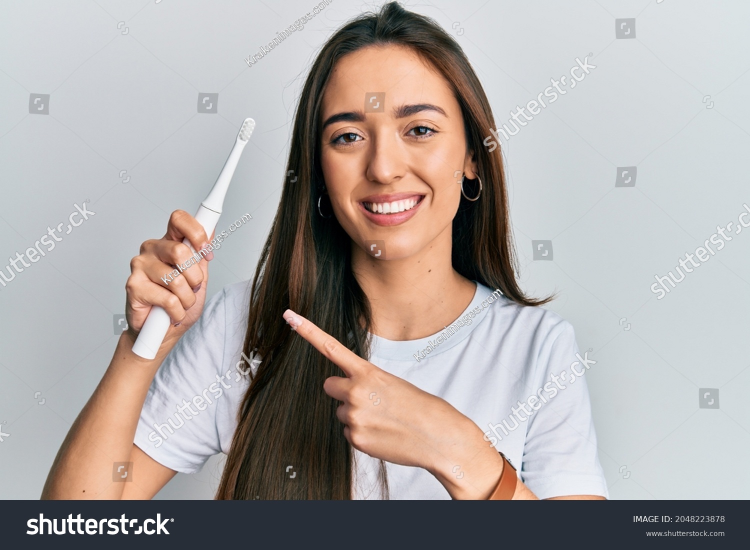 Young hispanic girl holding electric toothbrush smiling happy pointing with hand and finger  #2048223878