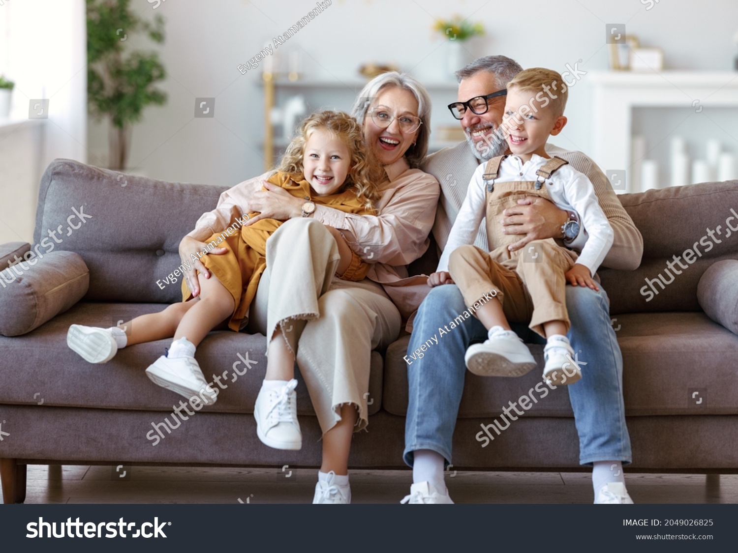 Happy family joyful little children hugging embracing with positive senior grandparents while sitting together on sofa in living room at home, cheerful grandma and grandpa with kids smiling at camera #2049026825