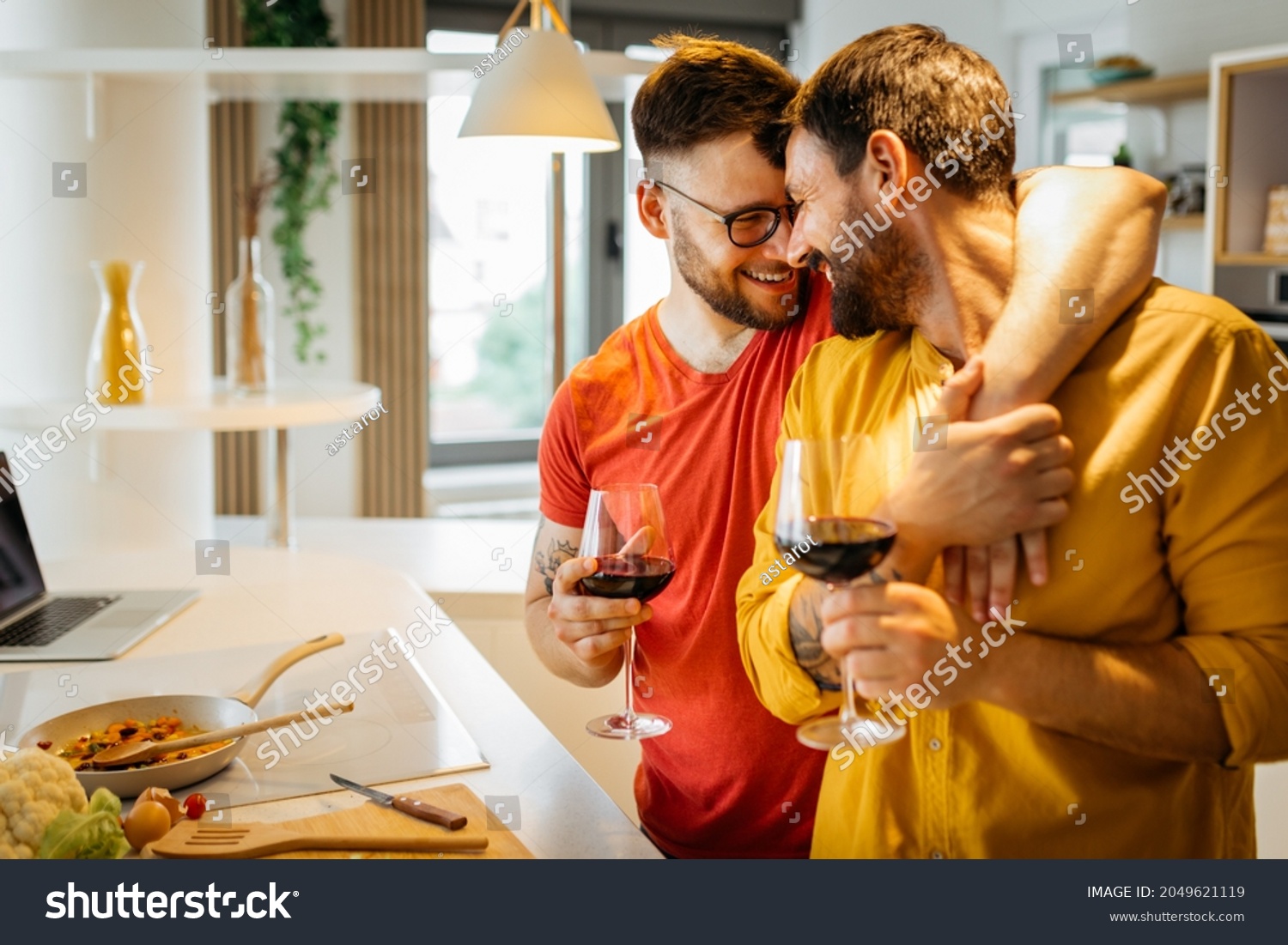 Happy hipster gay couple in love hugging while standing in the kitchen. They are cooking dinner together at home and drinking wine #2049621119