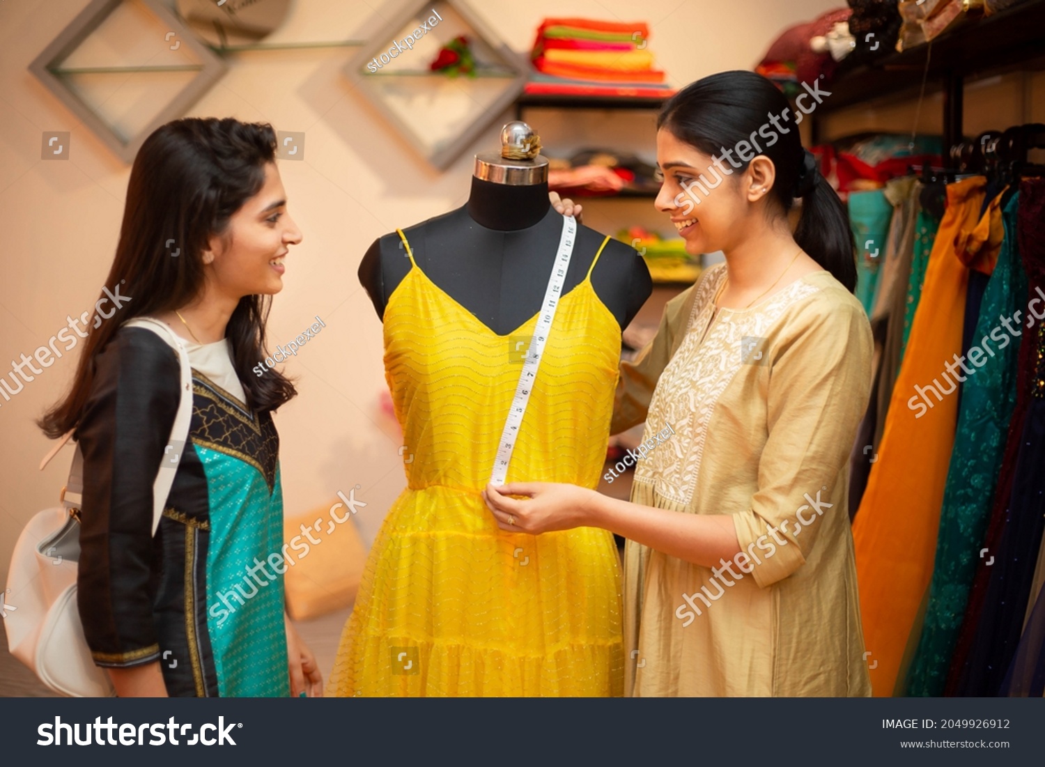 Young happy indian woman,tailor seamstress,smiling fashion designer showing customer or client dress measurements with tape,measuring on mannequin,standing in garment workshop,atelier or studio. #2049926912