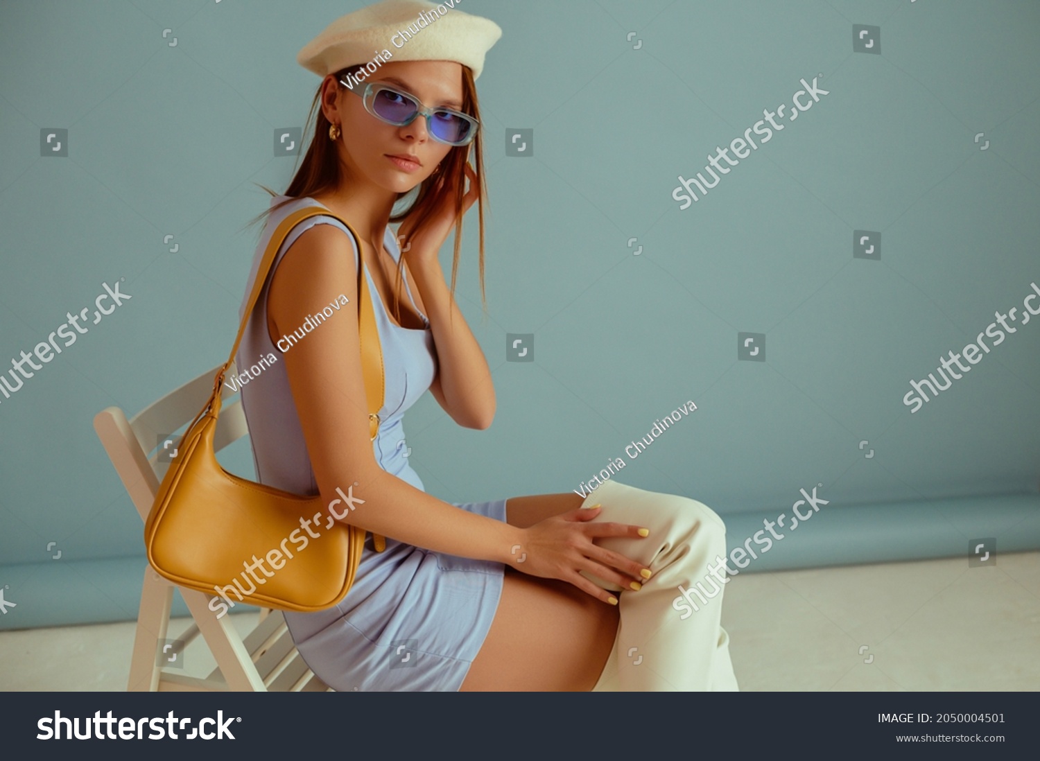 Fashionable woman wearing classic white beret, color sunglasses, elegant mini dress, over knee boots, with yellow leather shoulder bag, posing on blue background. Copy, empty space for text #2050004501