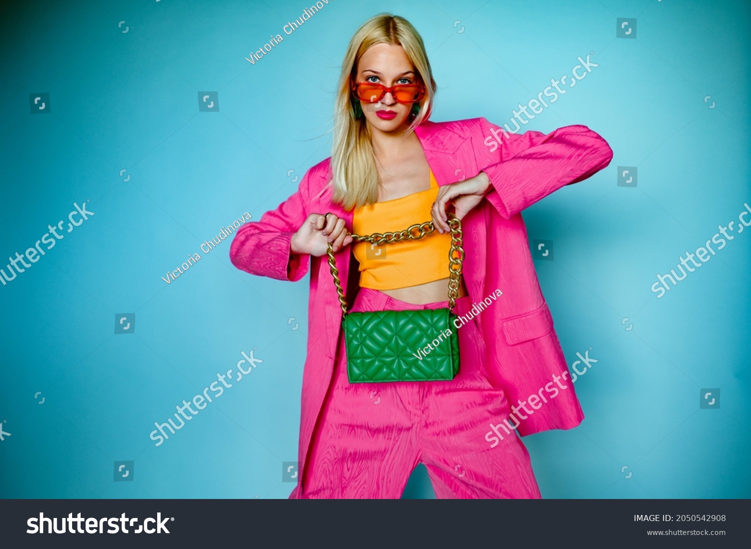 Fashion portrait of confident woman wearing trendy summer pink fuchsia color suit, orange sunglasses, holding green quilted faux leather bag, posing in studio, on blue background. Copy, empty space
 #2050542908