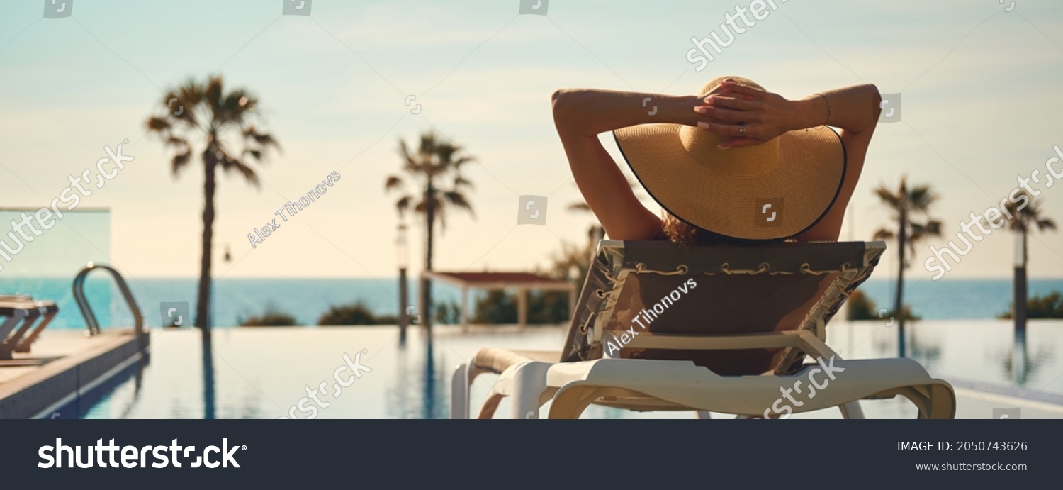 Rear view woman wear hat lying on deckchair near pool, put hands behind head relaxing, take sun bath, sea palm tree empty swimming pool scenery on background. Summer holidays, vacation, travel concept #2050743626