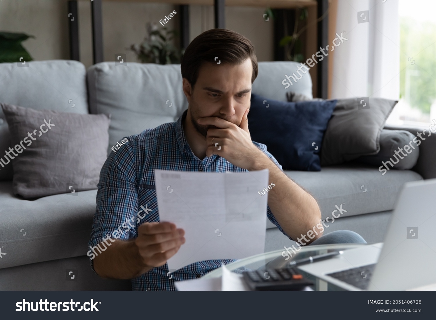 Thoughtful man checking financial documents, holding receipt, calculating domestic bills or taxes, using laptop, unhappy pensive young male touching chin, unexpected debt or bankruptcy concept #2051406728
