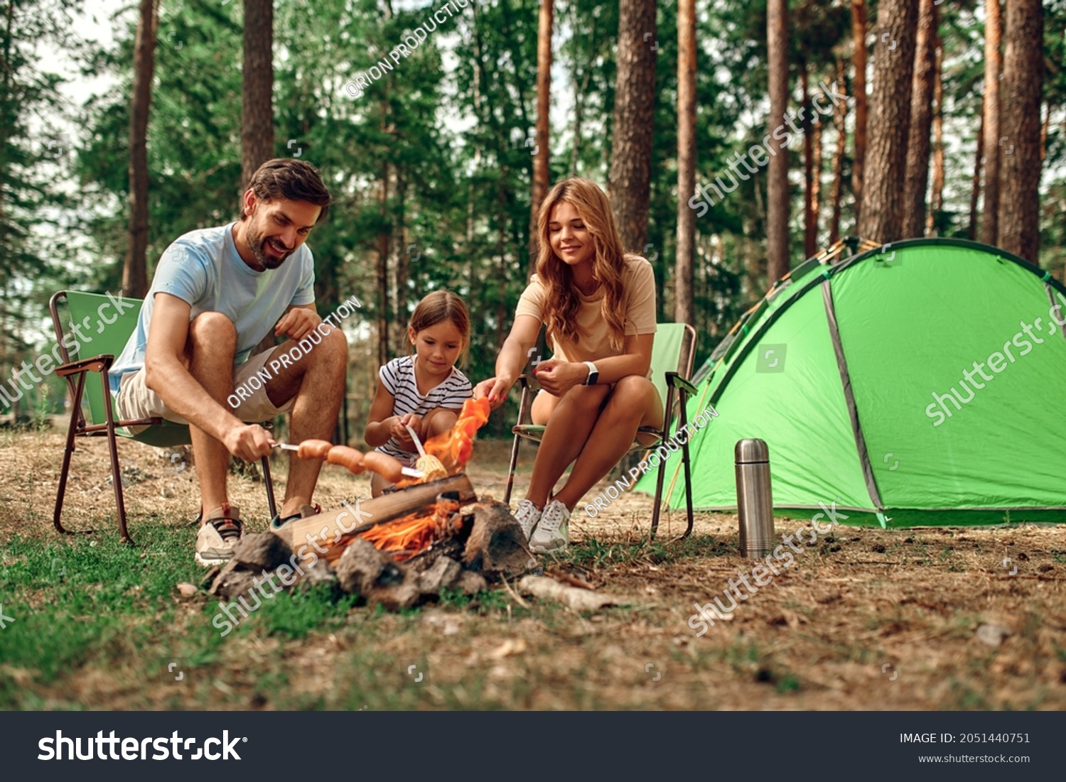 Happy family with a child on a picnic sit by the fire near the tent and grill a barbecue in a pine forest. Camping, recreation, hiking. #2051440751