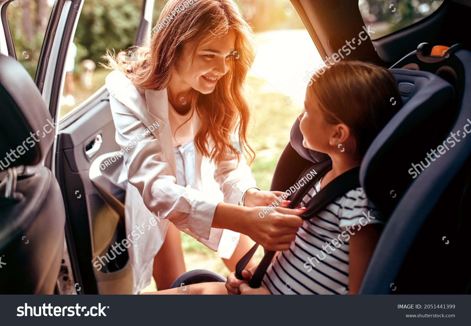 A cute woman mother put her daughter in a car seat and fastens her seat belts. Protection during the trip in the car. #2051441399