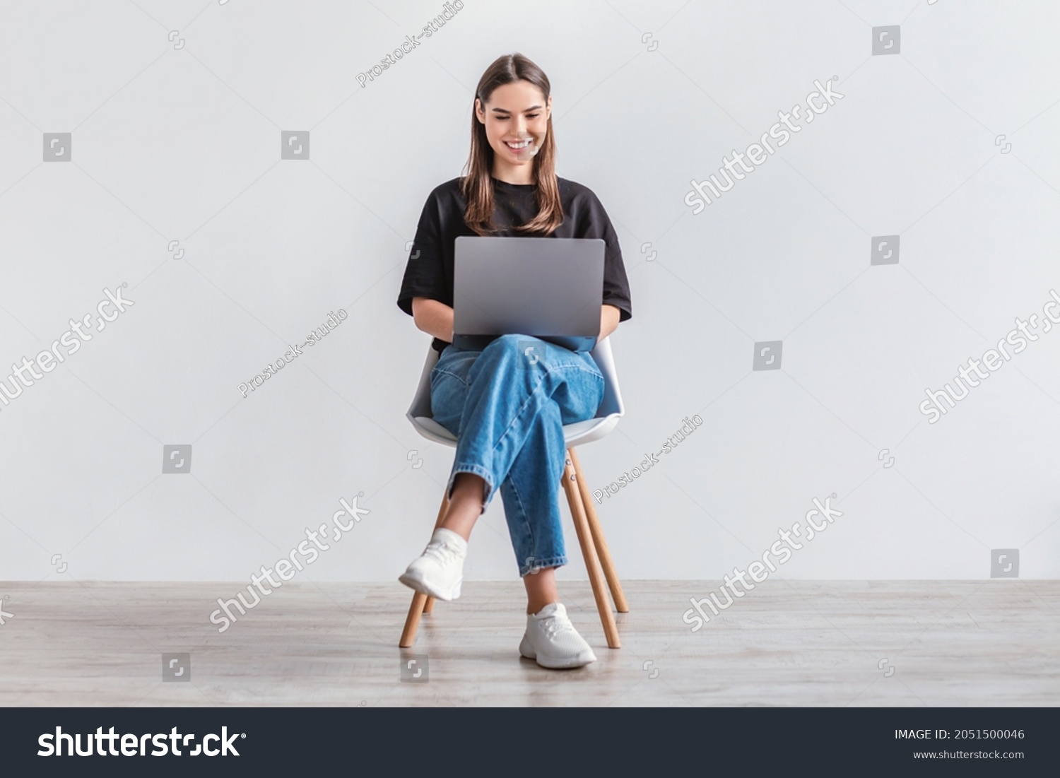 Happy young woman working online, sitting on chair and using laptop against white studio wall, full length. Positive Caucasian lady surfing internet, watching webinar on portable computer #2051500046
