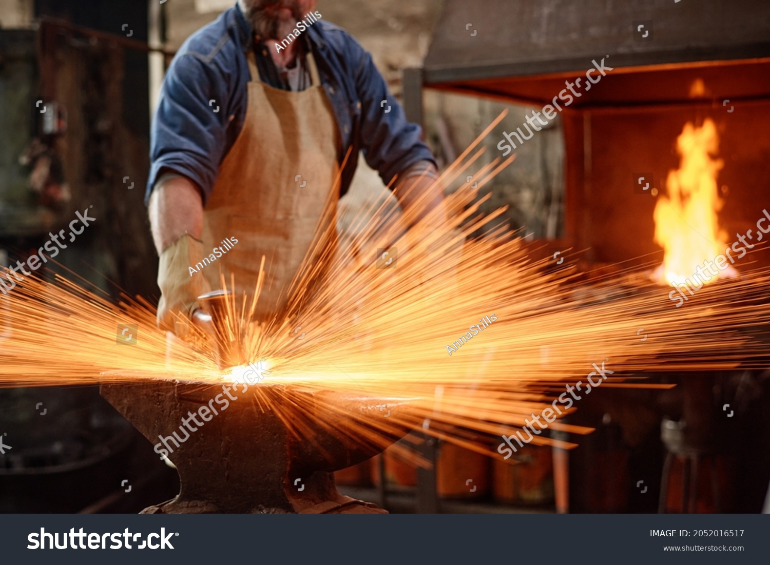 Blacksmith in apron forging the iron with hammer while working in the workshop #2052016517