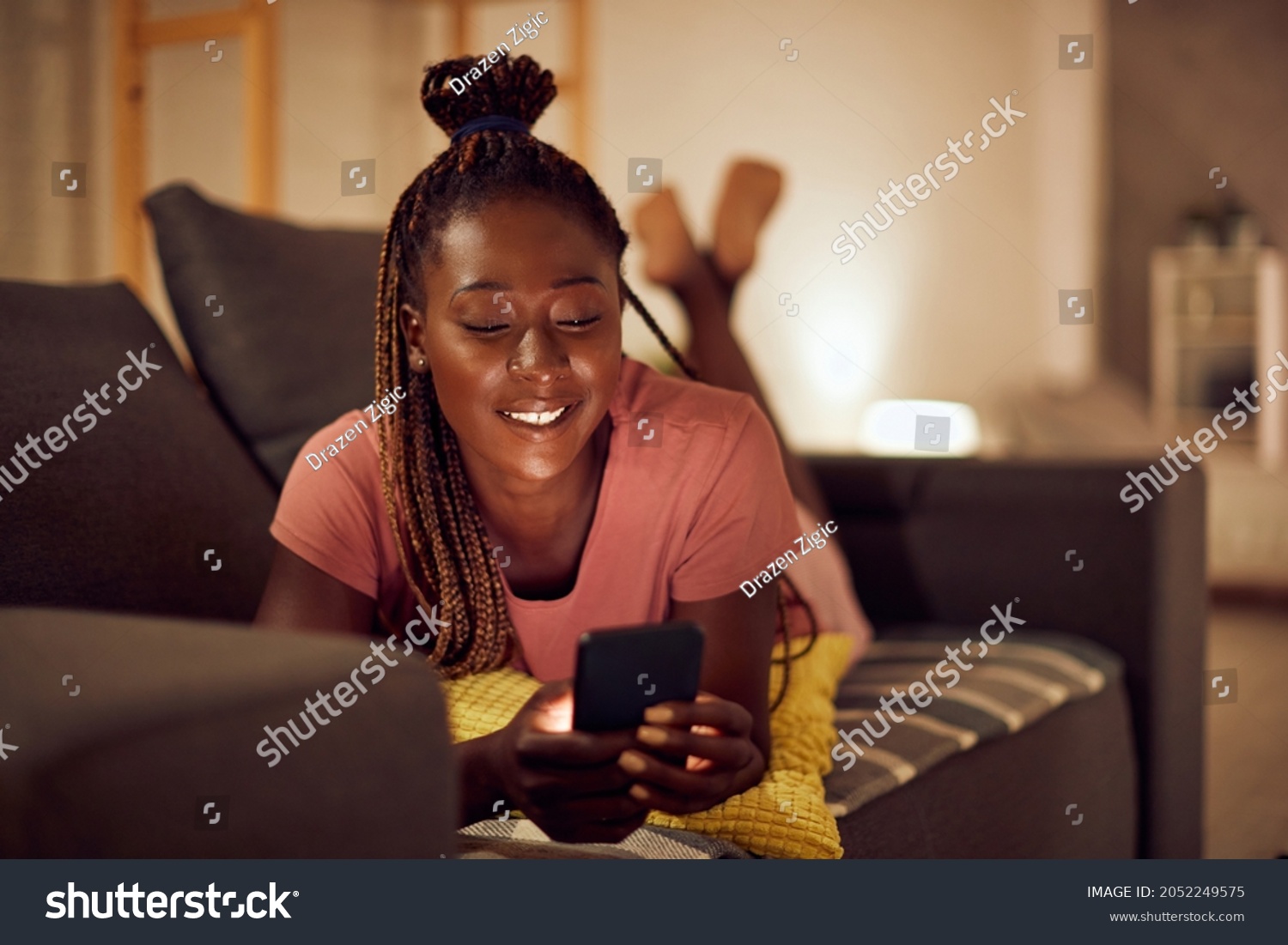 Happy black woman using smart phone and text messaging while relaxing at night in the living room.  #2052249575